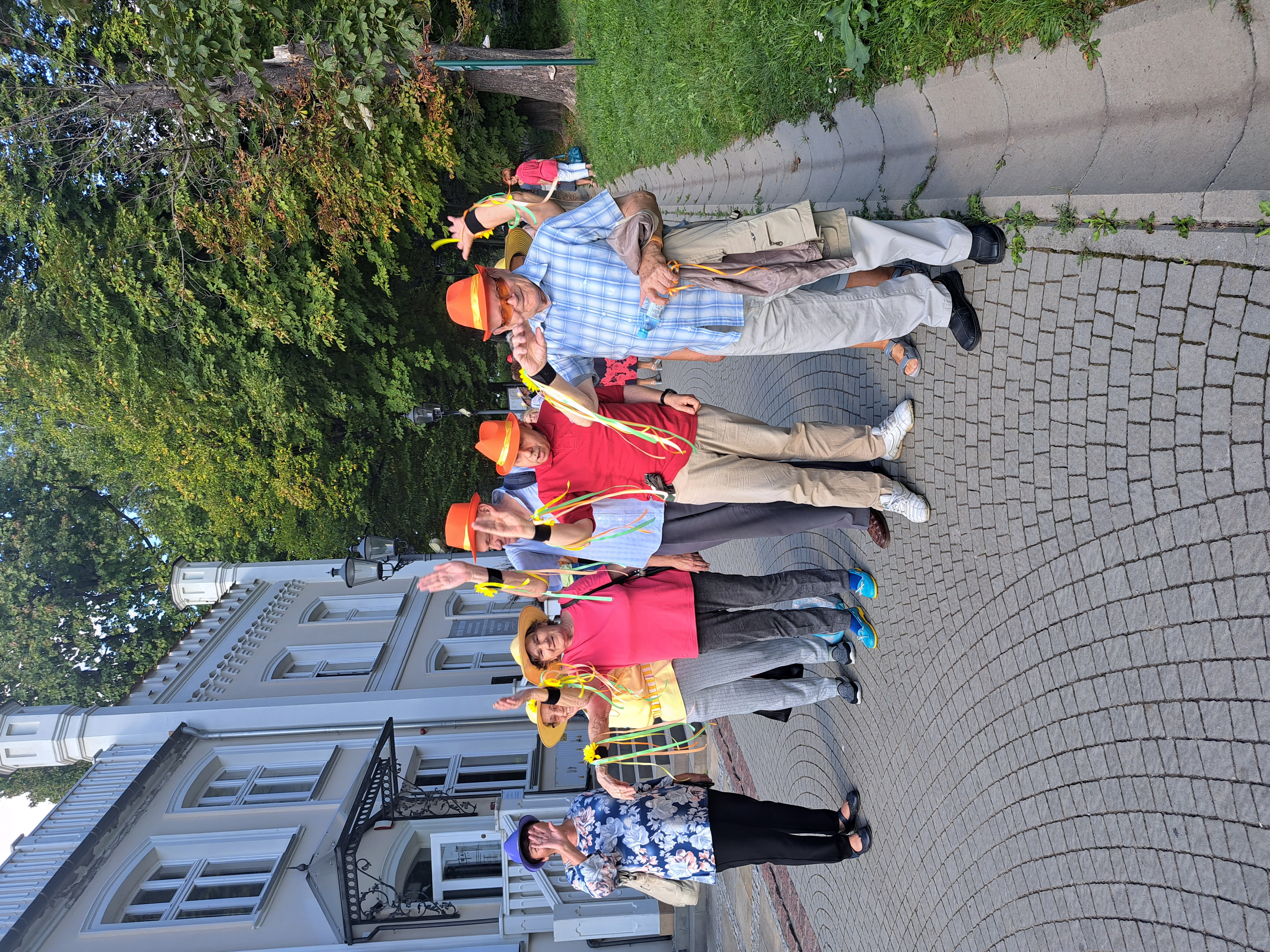 Grupa Seniorów pozuje w Parku Strzeleckim obok budynku BWA. Na głowach mają czerwone kapelusze