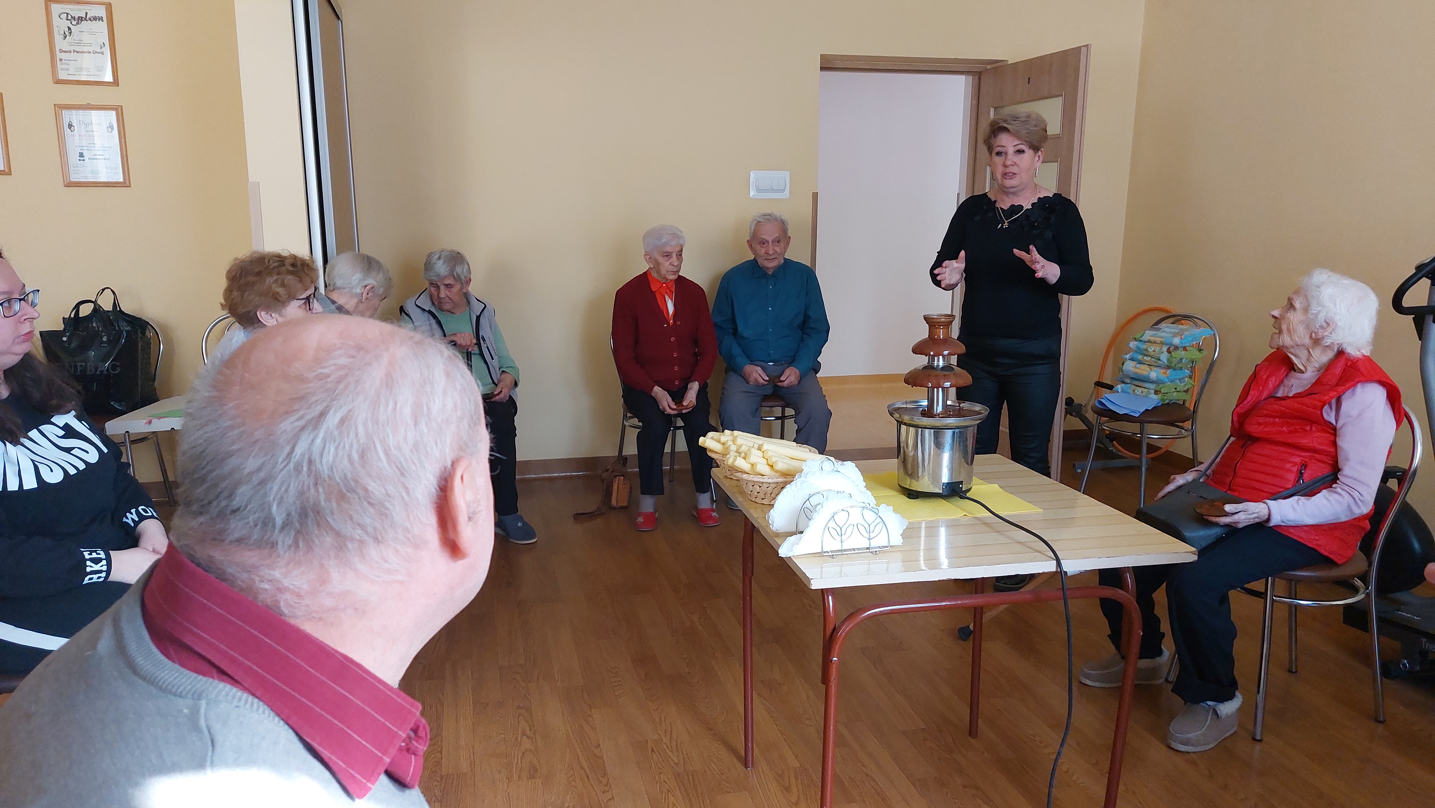 Na środku sali stoi stolik, na nim znajduje się fontanna czekoladowa. Dokoła siedzą Seniorzy.