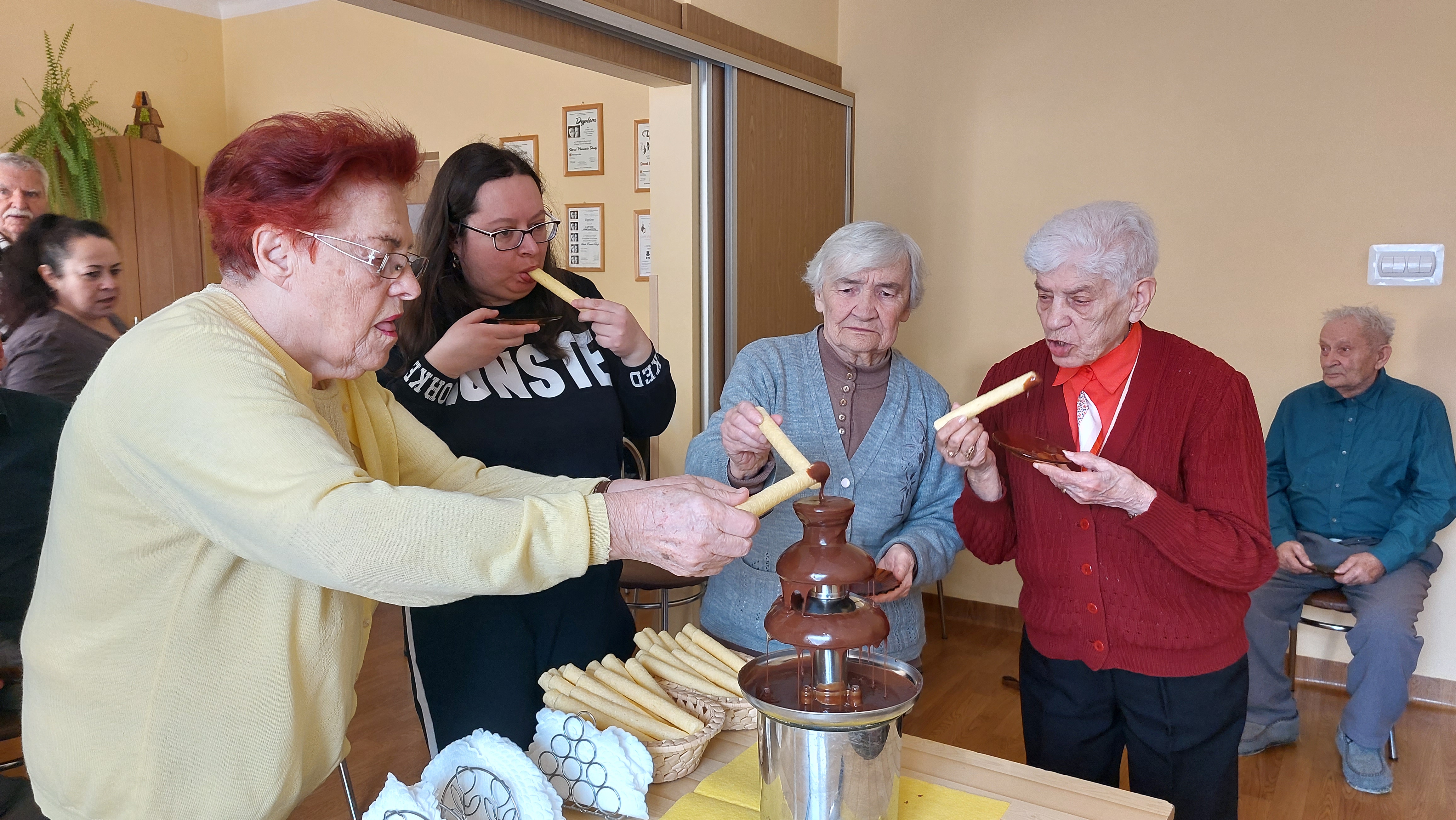 Grupa Seniorów stoi przy fontannie czekoladowej, maczają chrupki w czekoladzie