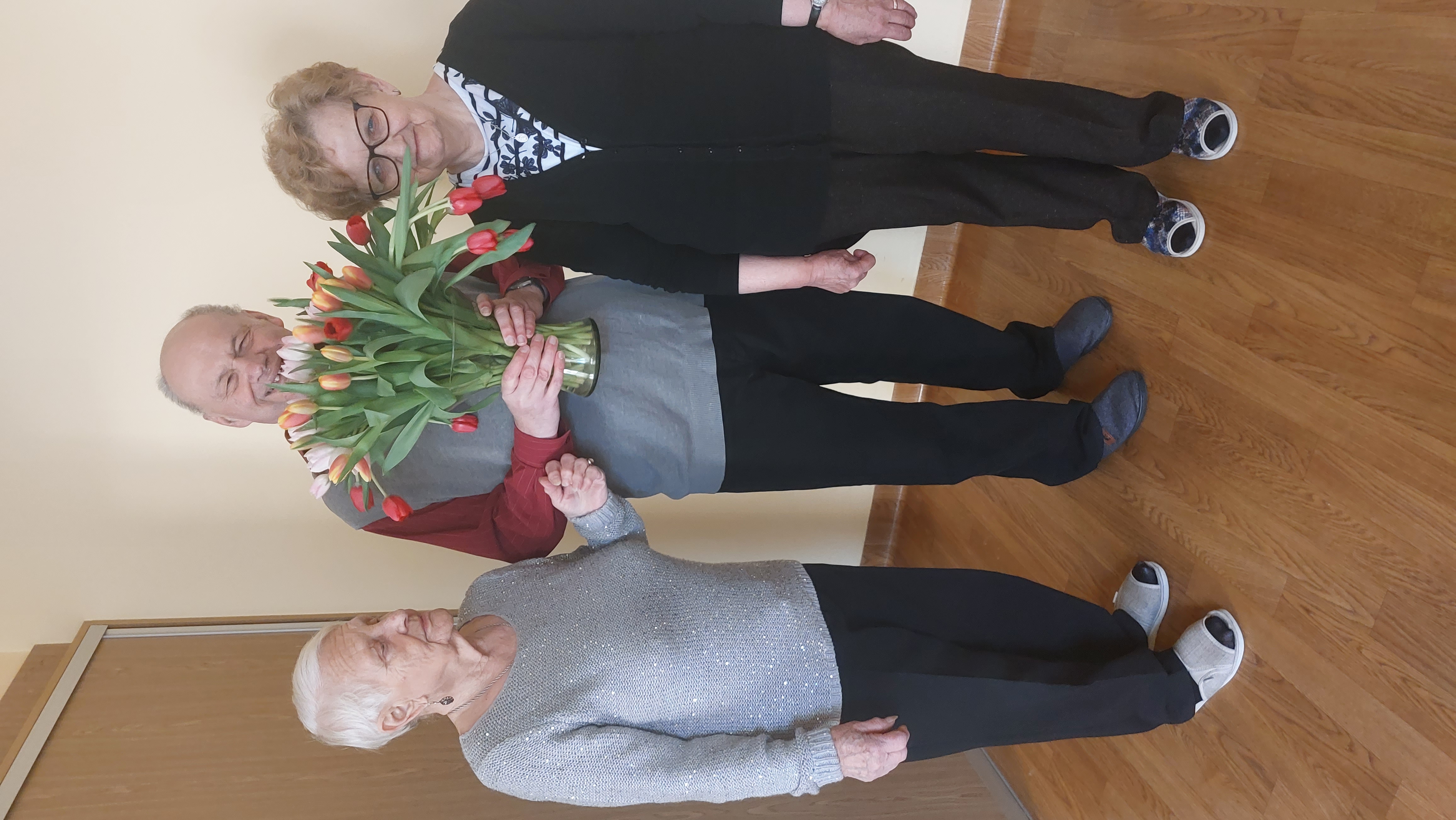 Senior trzymająby bukiet tulipanów, po jego prawej oraz lewej stronie stoją dwie Seniorki