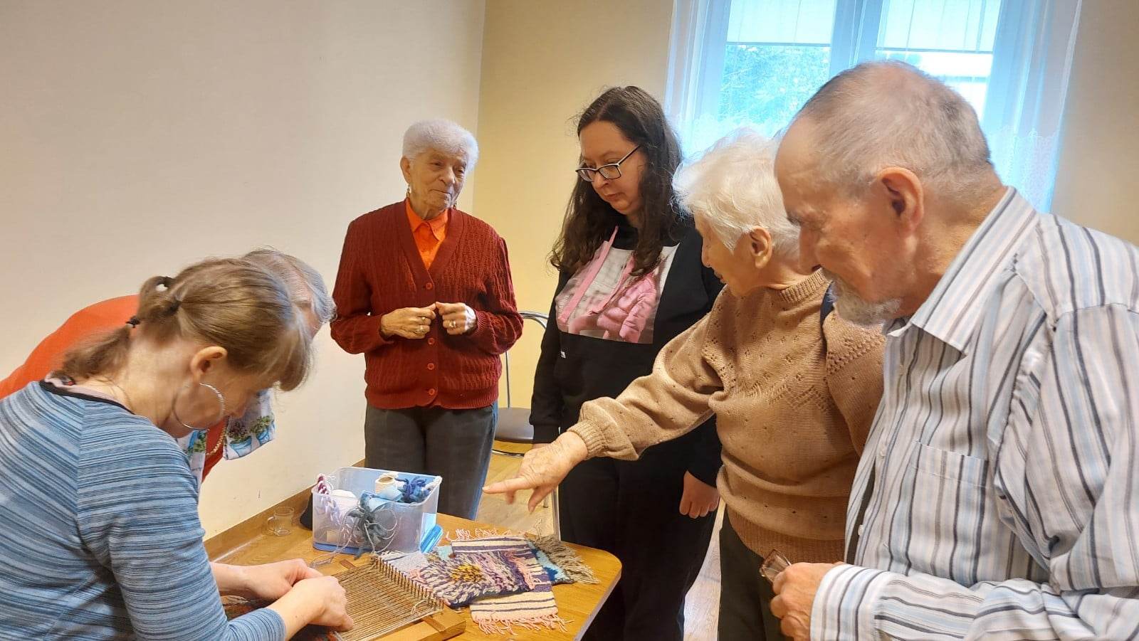 Seniorka pochyla się, by porozmawiać z Panią Anią. Dokoła stoją inni Seniorzy