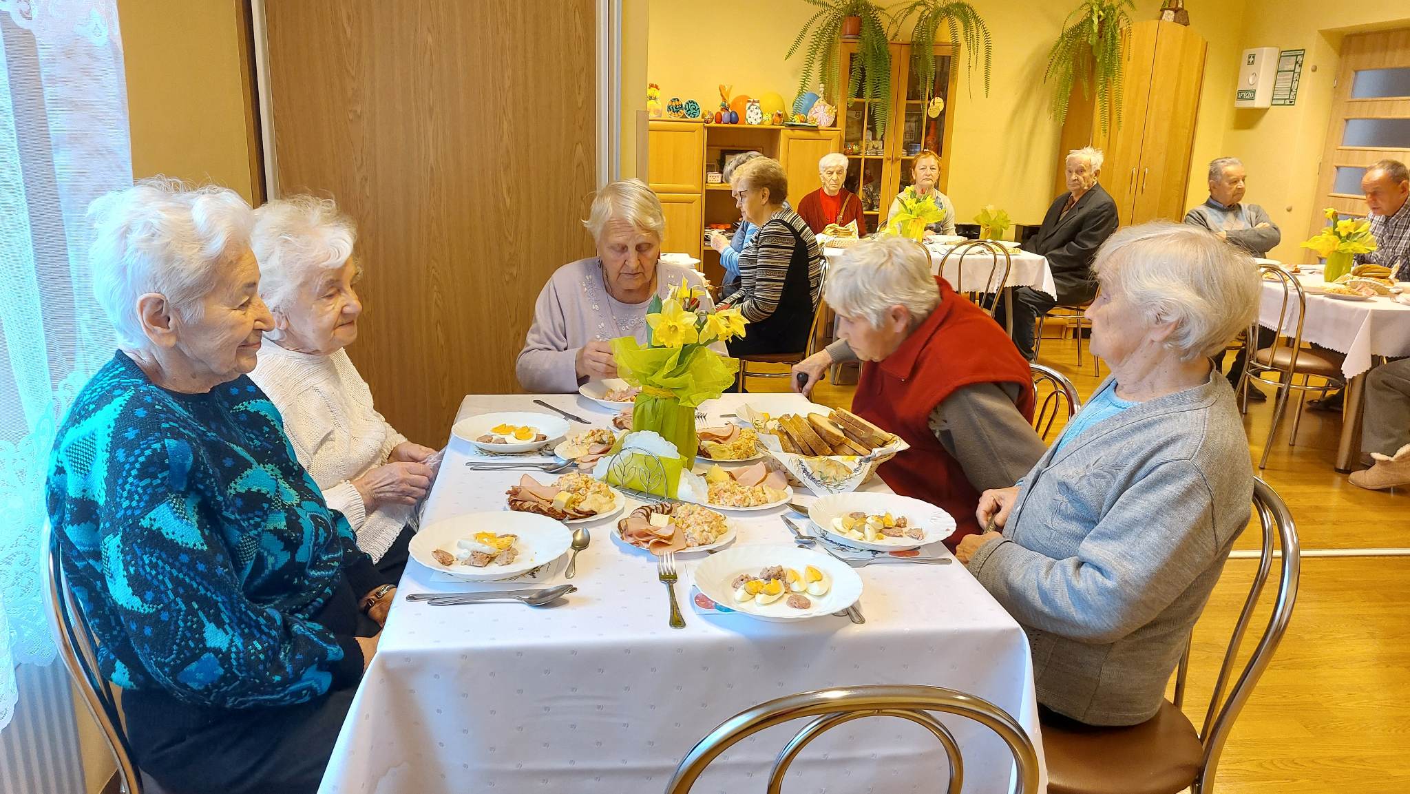 Seniorzy siedzą przy stole, zastawionym  sałatkami warzywnymi, wędlinami, jajkami