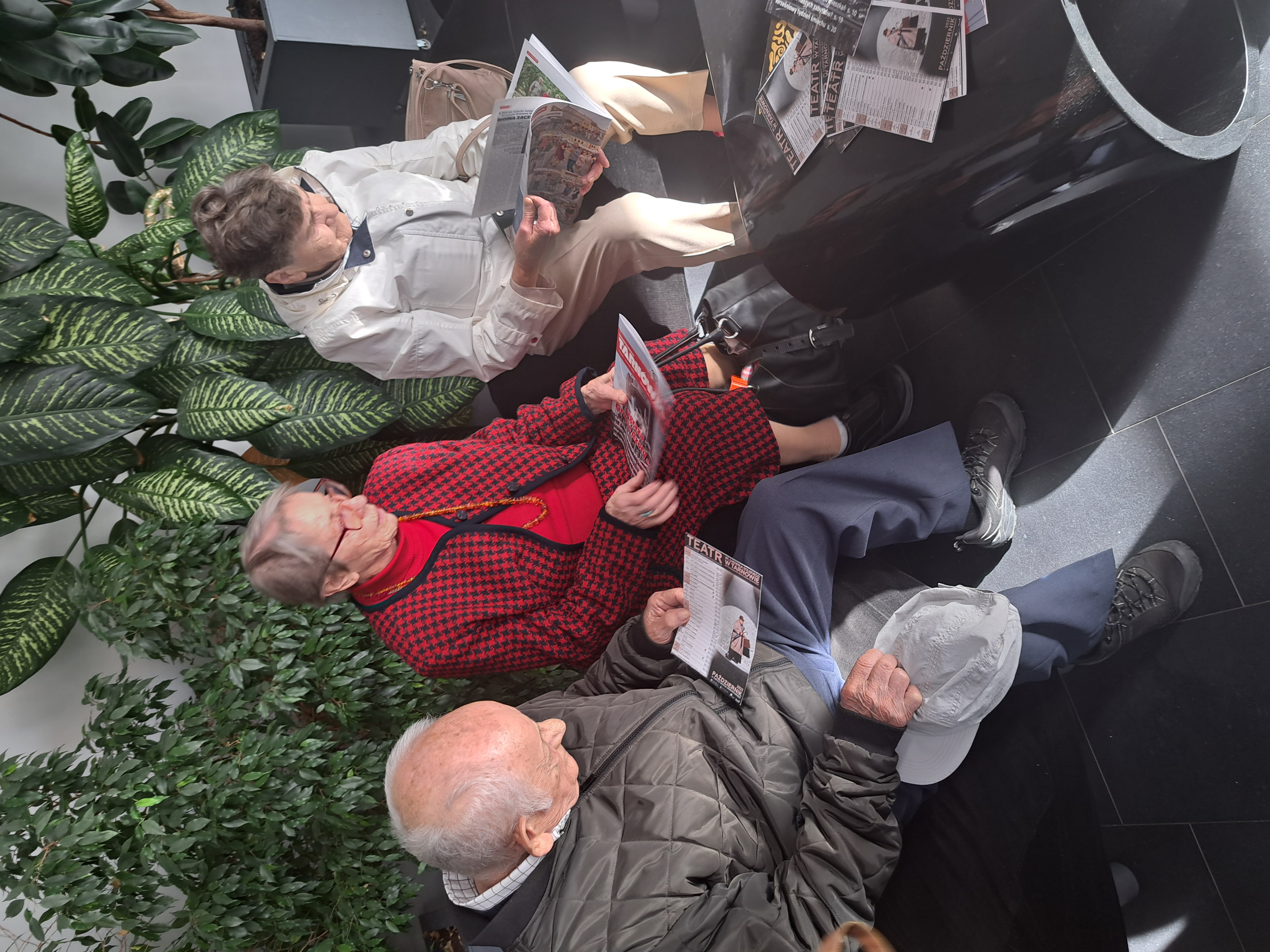 Troje Seniorów siedzi na kanapie w budynku teatru, przeglądają gazetki miejskie