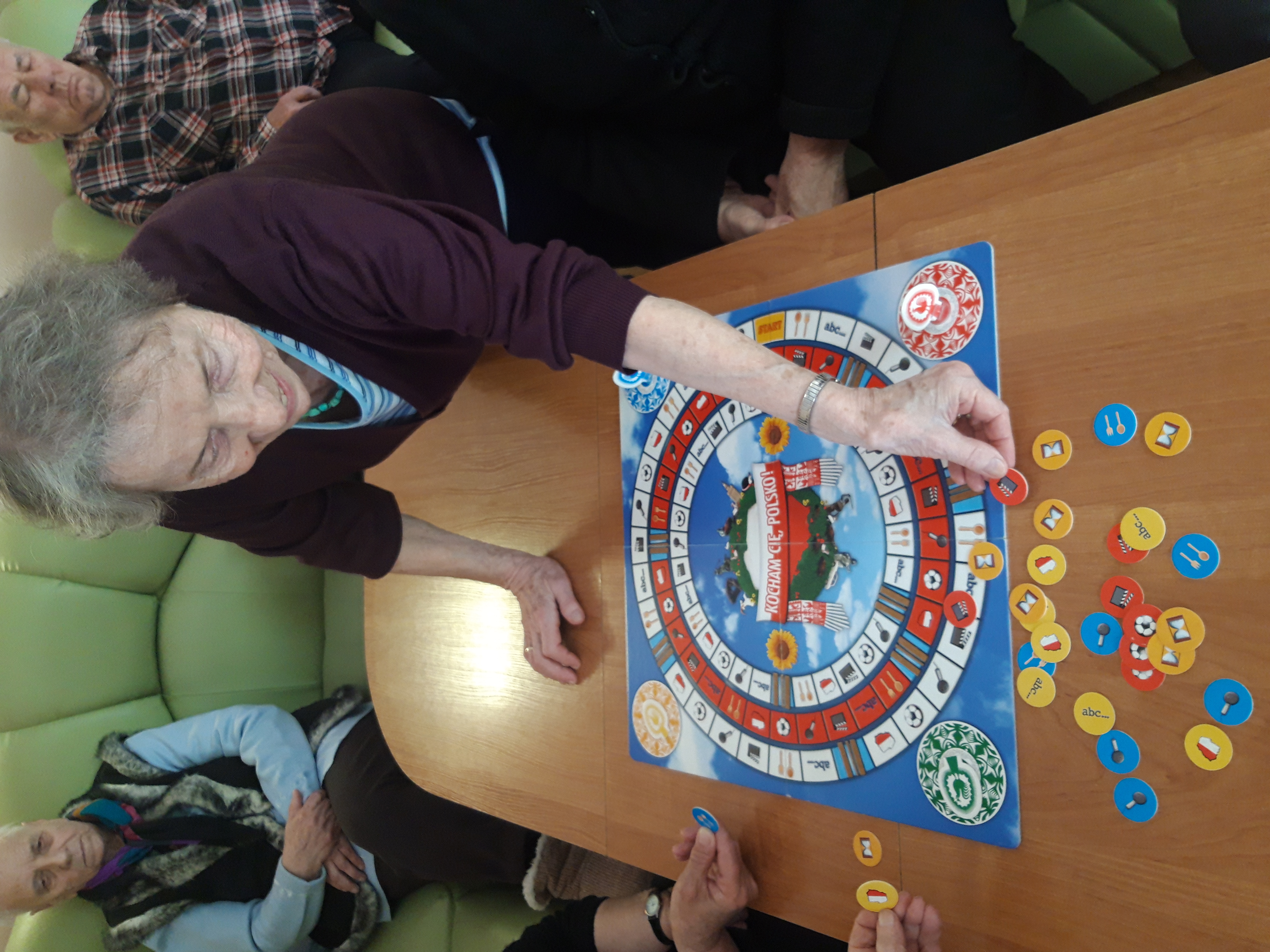 Seniorka opiera się ręką na stole, na którym leży gra planszowa, w drugiej dłoni trzyma żeton do gry