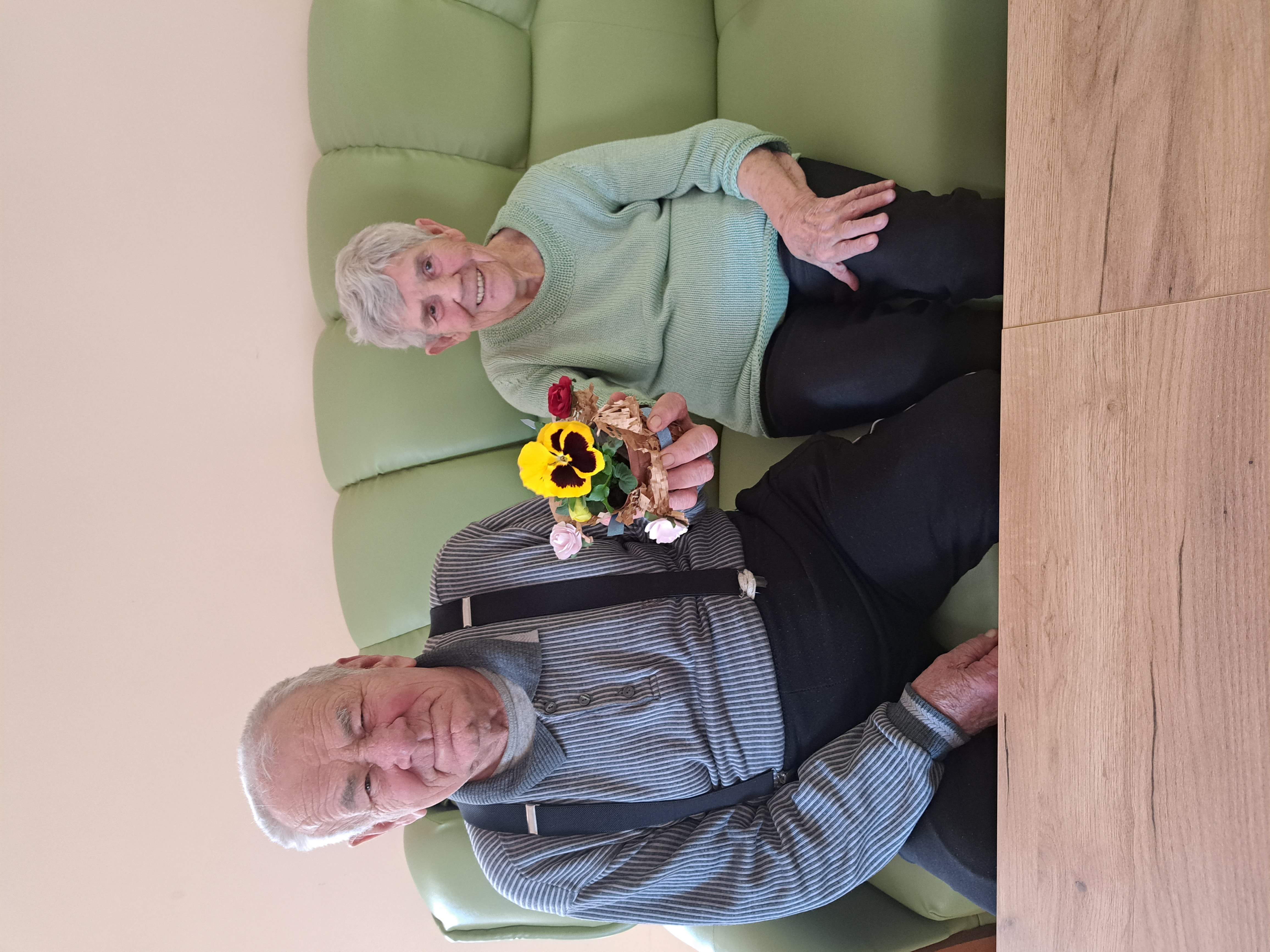 Seniorka wraz z Seniorem siedzi na kanapie, w wyciągniętej przed siebie dłoni trzyma żółtego kwiatka w doniczce