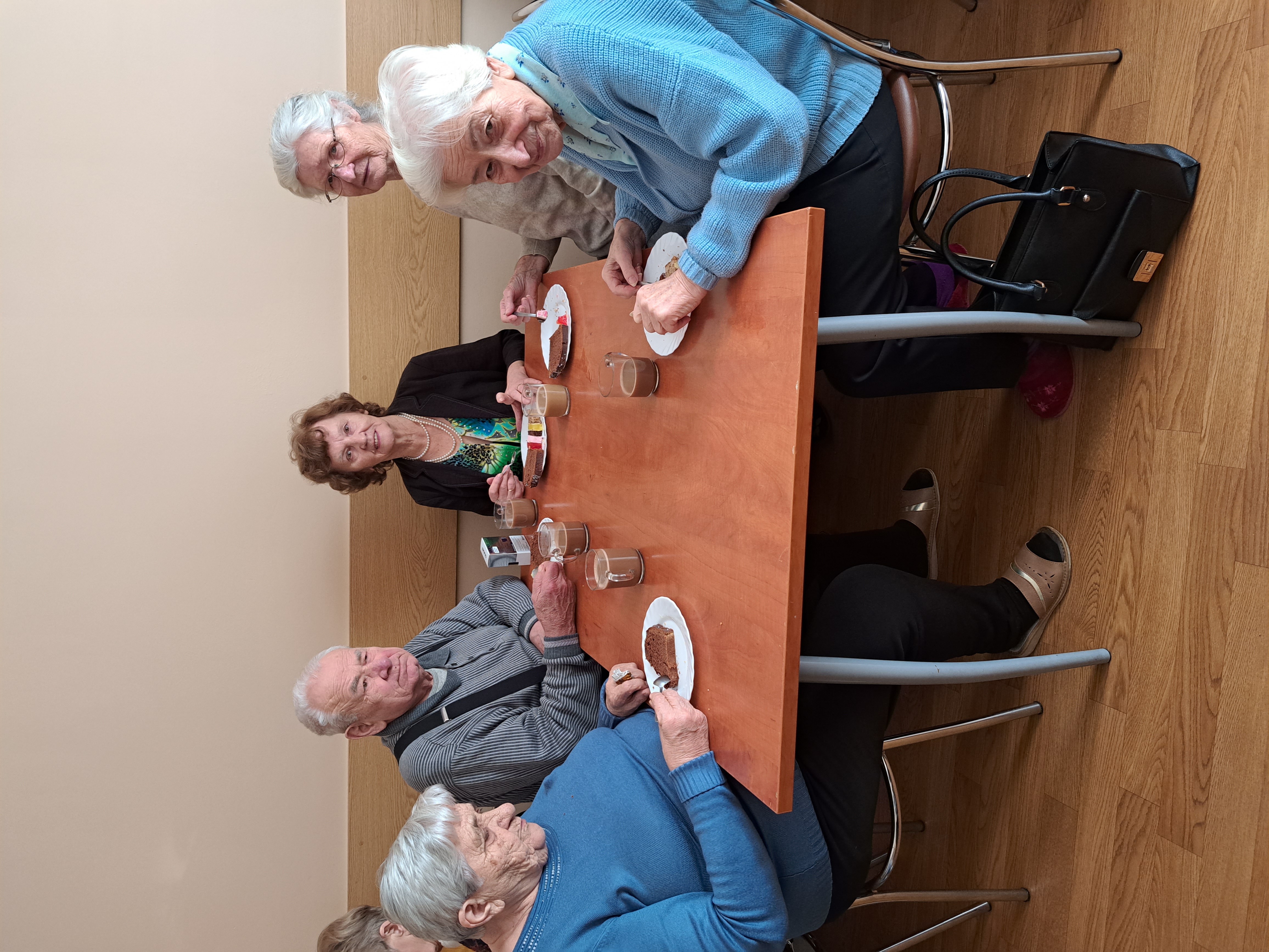 Seniorzy siedzą przy stoliku, każdy z nich ma ciastko oraz kawę