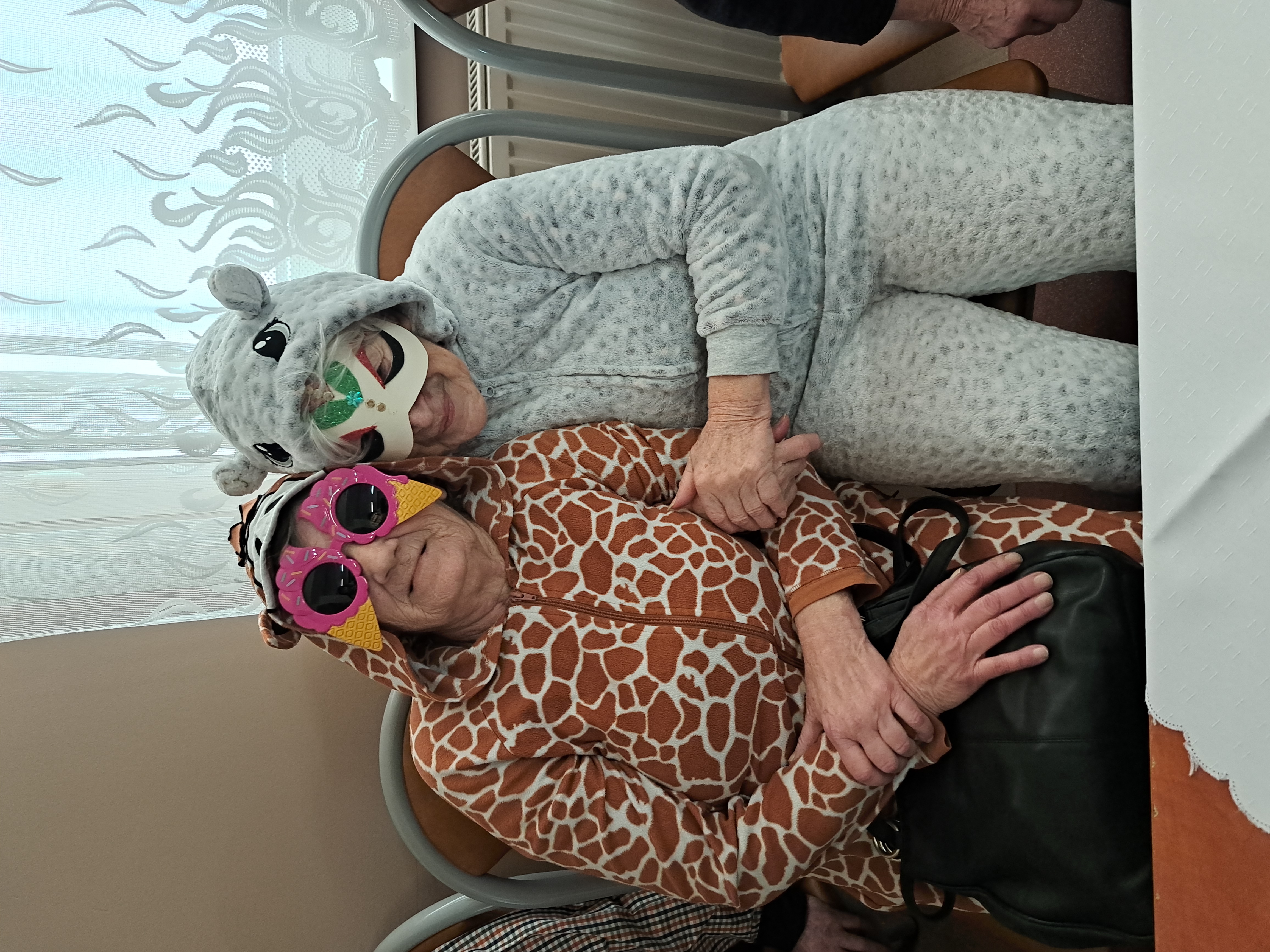 Dwie Seniorki, ubrane w bluzy z motywem zwierzęcym i maskami na twarzach, siedzą na kanapie i obejmują się do zdjęcia