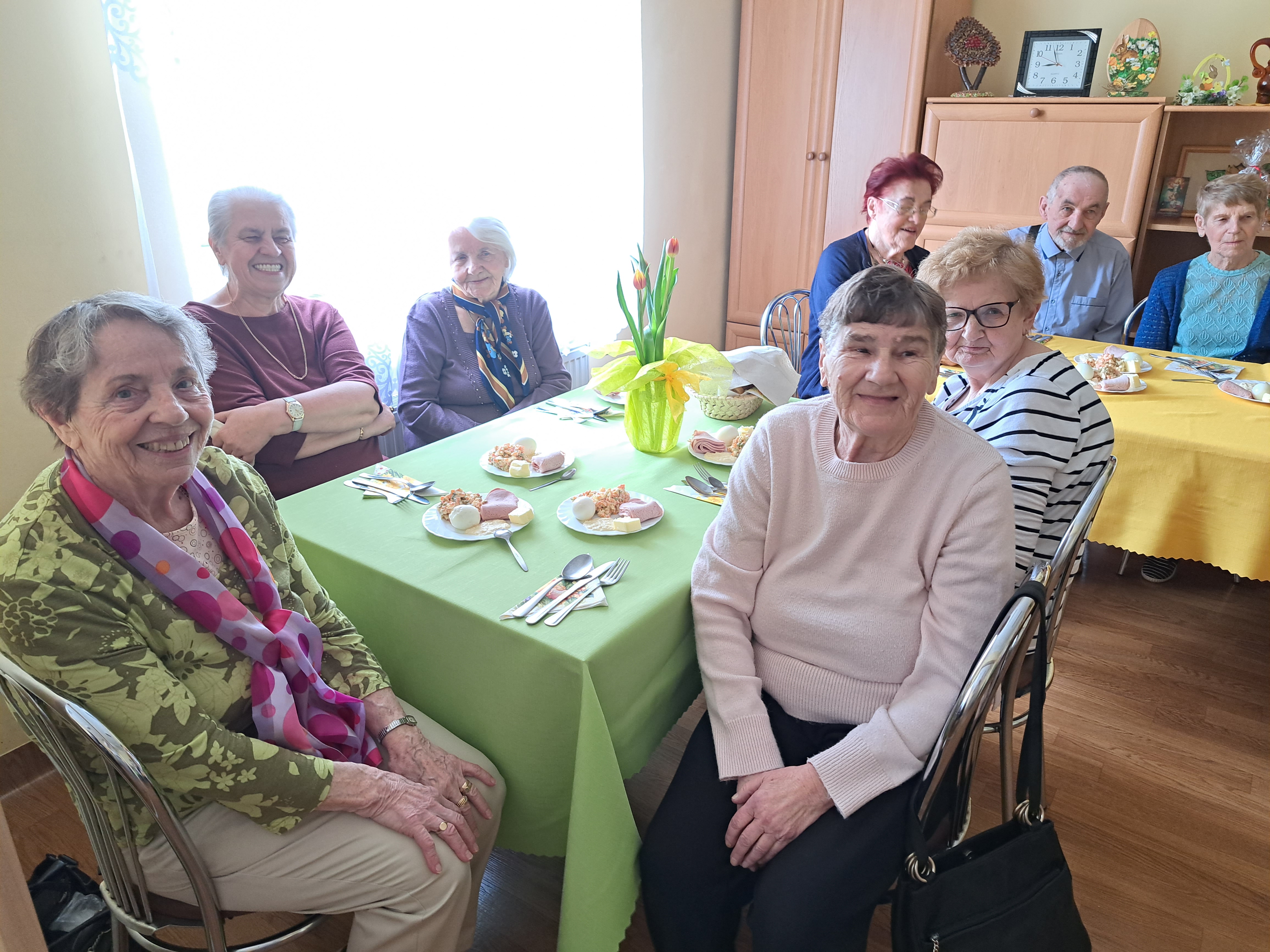 Seniorzy siedzą przy zastawionych wielkanocnych stołach, uśmiechają się do aparatu