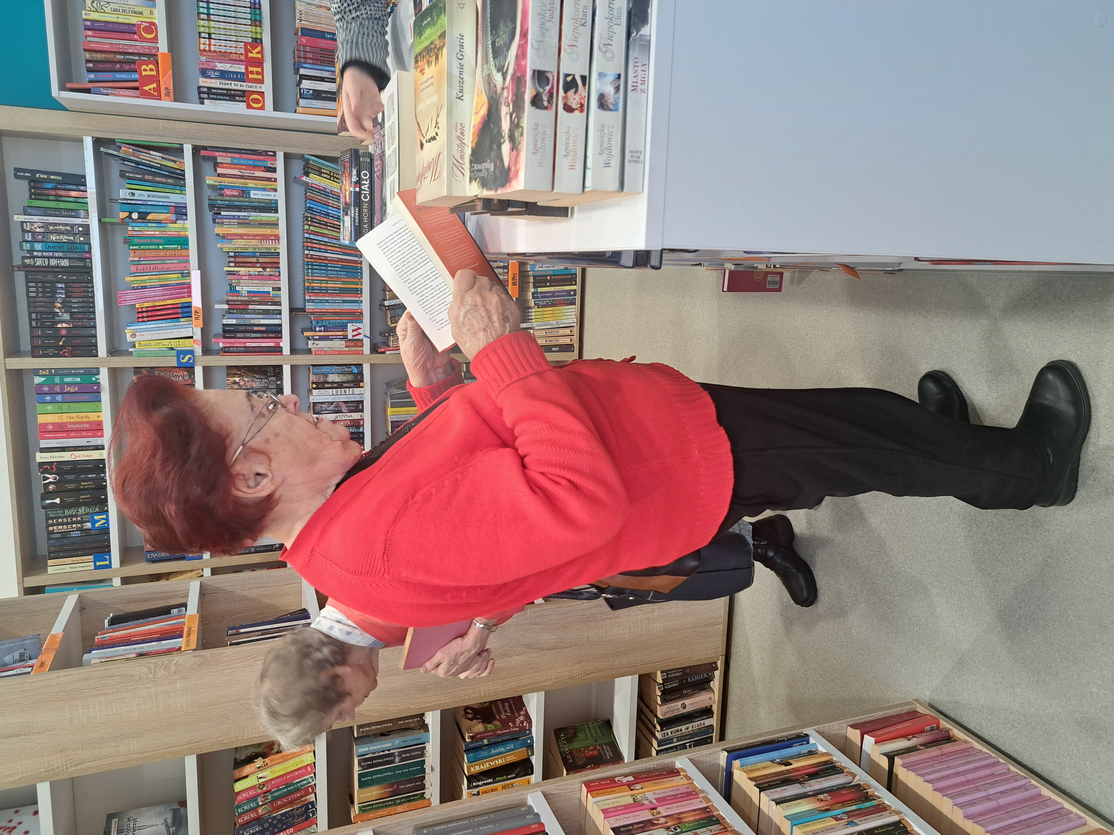 Seniorka stoi przy regale z książkami, jedną z książek przegląda
