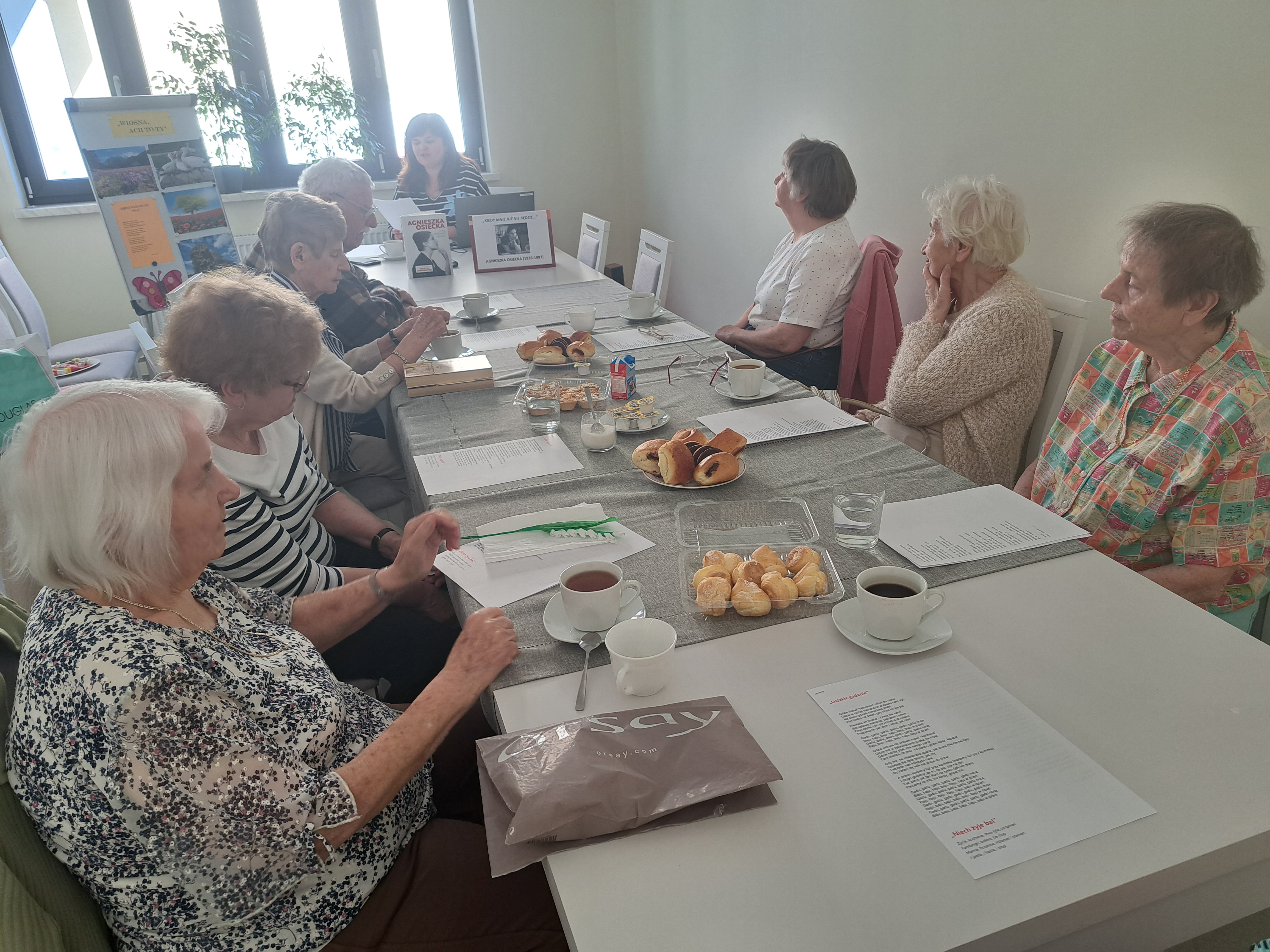 Seniorzy siedzą przy stole, rozmawiają z Panią prowadzącą zajęcia z biblioterapii