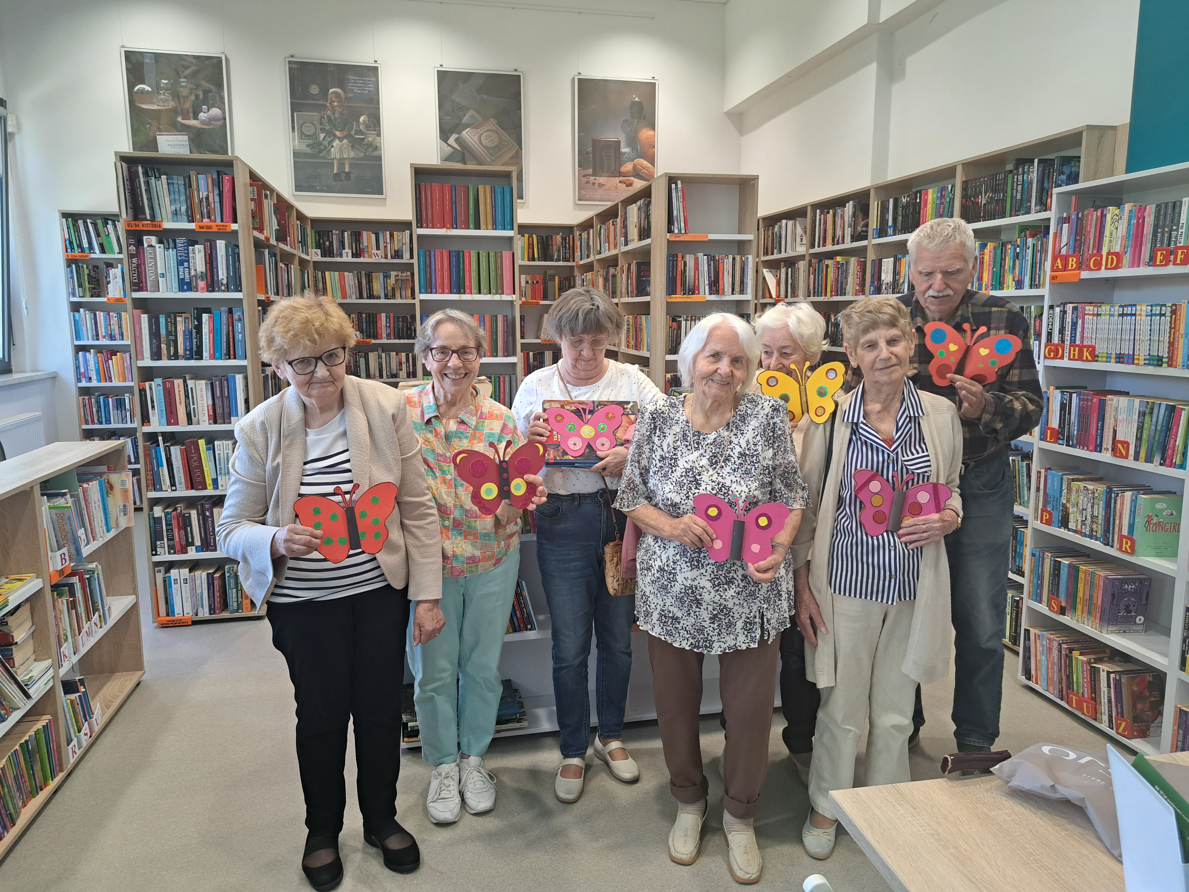 Seniorzy, na tle biblioteczek, pozują z własnoręcznie wykonanymi motylami z papieru