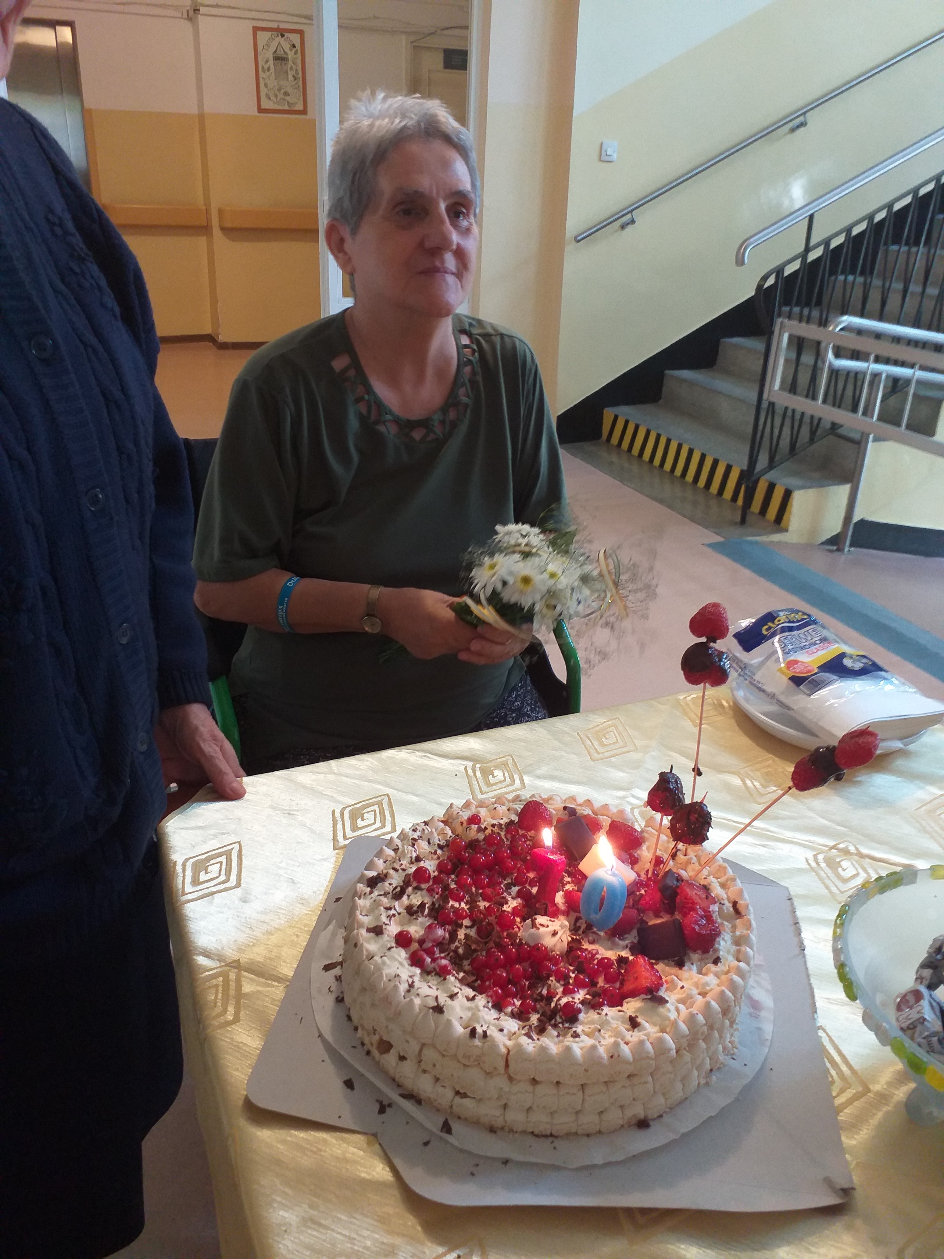 Seniorka siedzi przy stole, przed nią stoi tort urodzinowy