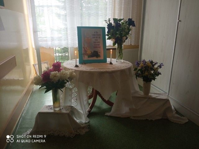 Okrągły stół, na którym znajdują się kwiaty, dwie zapalone świece oraz tabliczka z sentencją(nieczytelne)
