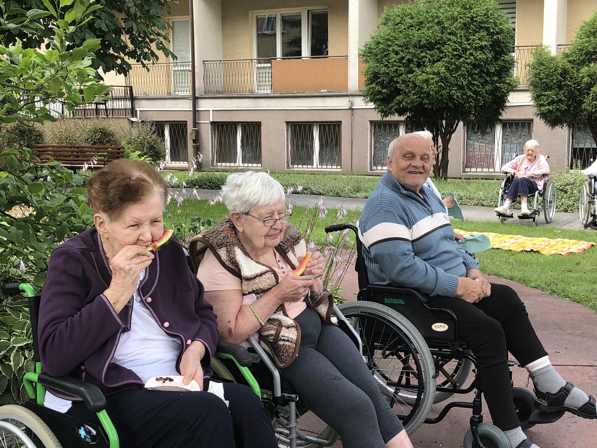 dwie Seniorki siedzą na wózkach, jedzą Seniora, na dalszym planie Senior uśmiecha się do zdjęcia