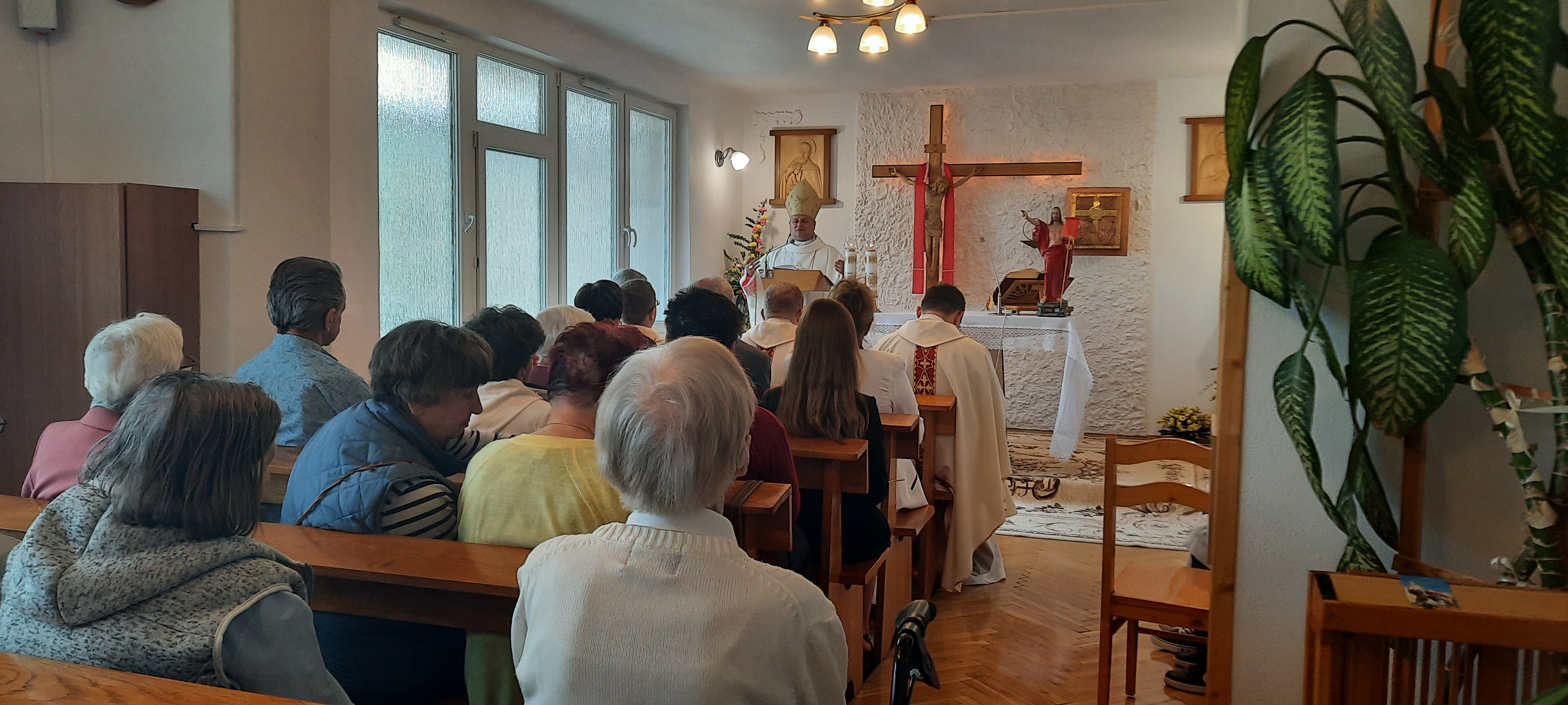 Na pierwszym planie ludzie siedzący w ławkach w kaplicy, z tyłu kapelan prowadzi nabożeństwo