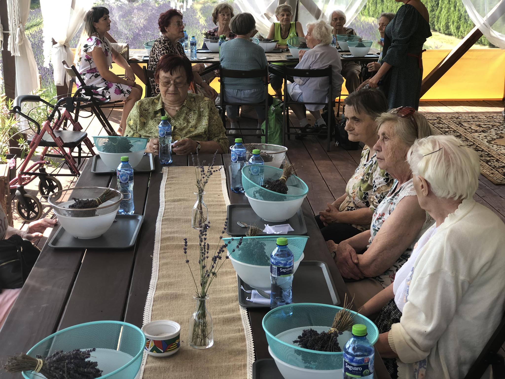 Seniorzy siedzą przy stole, przed każdym stoi miska z sitkiem, w którym znajduje się bukiet z kwiatów lawendy