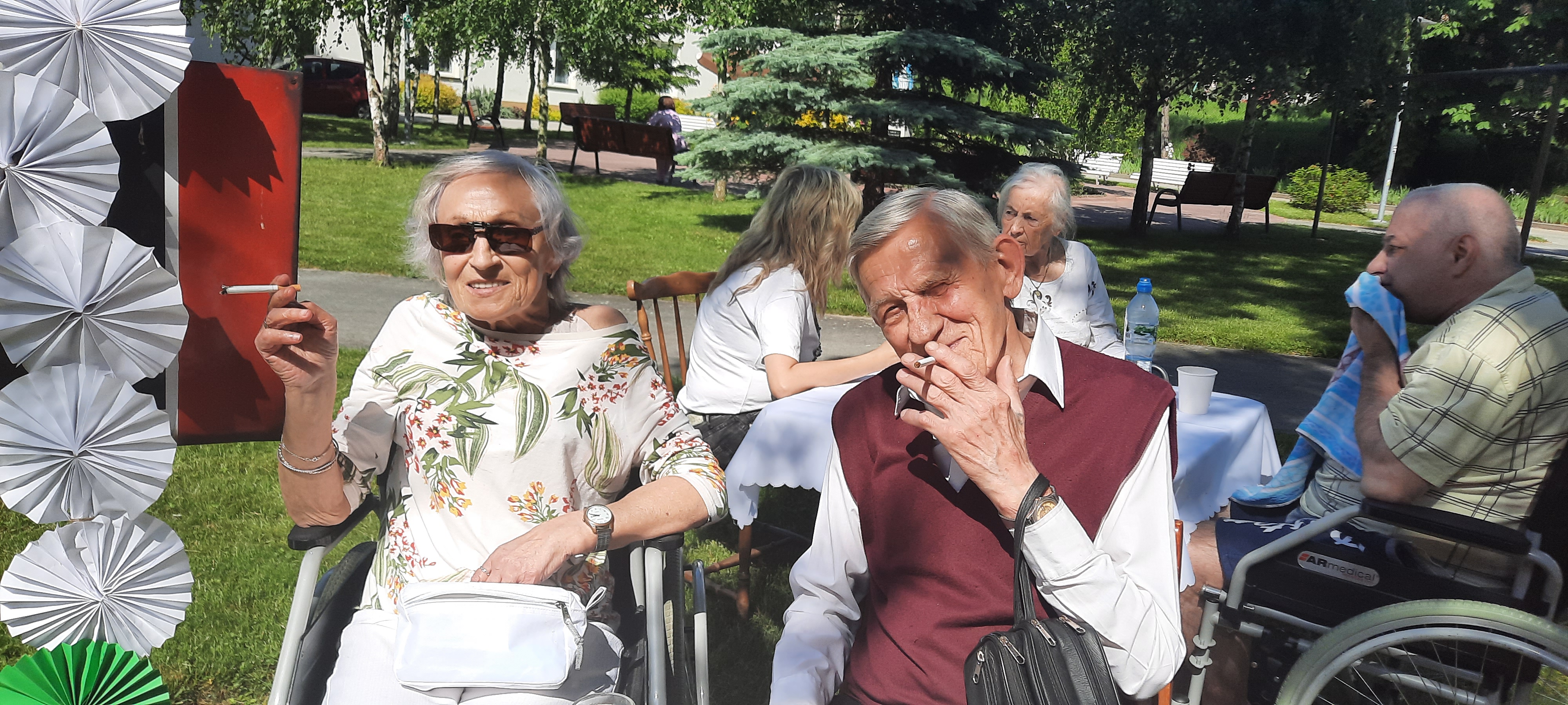 Senior i Seniorka siedzą w ogrodzie na wózkach inwalidzkich, uśmiechają się do obiektywu