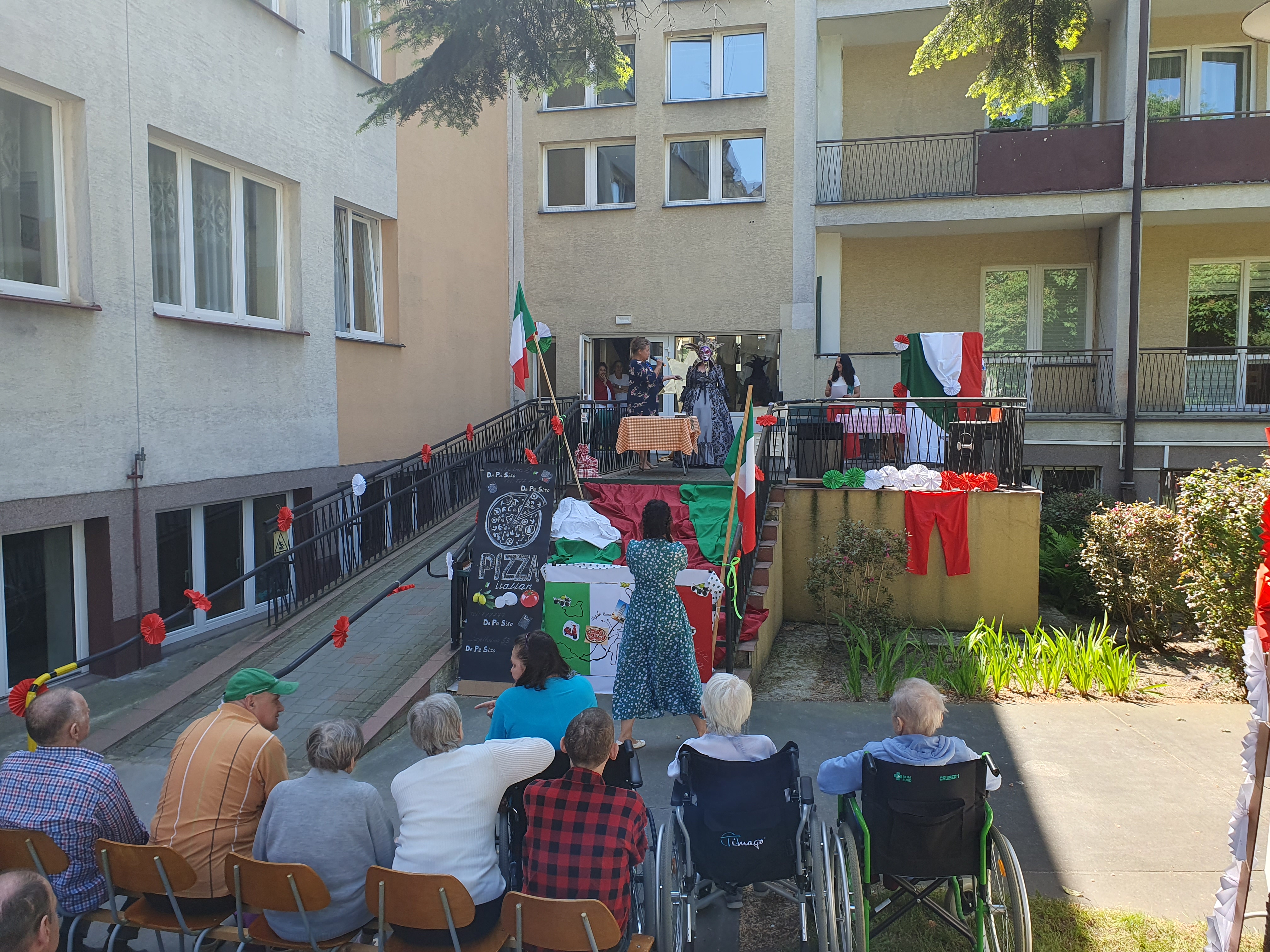 Na pierwszym planie znajdują się Seniorzy siedzący na ogrodzie, przed nimi znajdują się schody i wjazd do DPS zaaranżowane we włoskim stylu