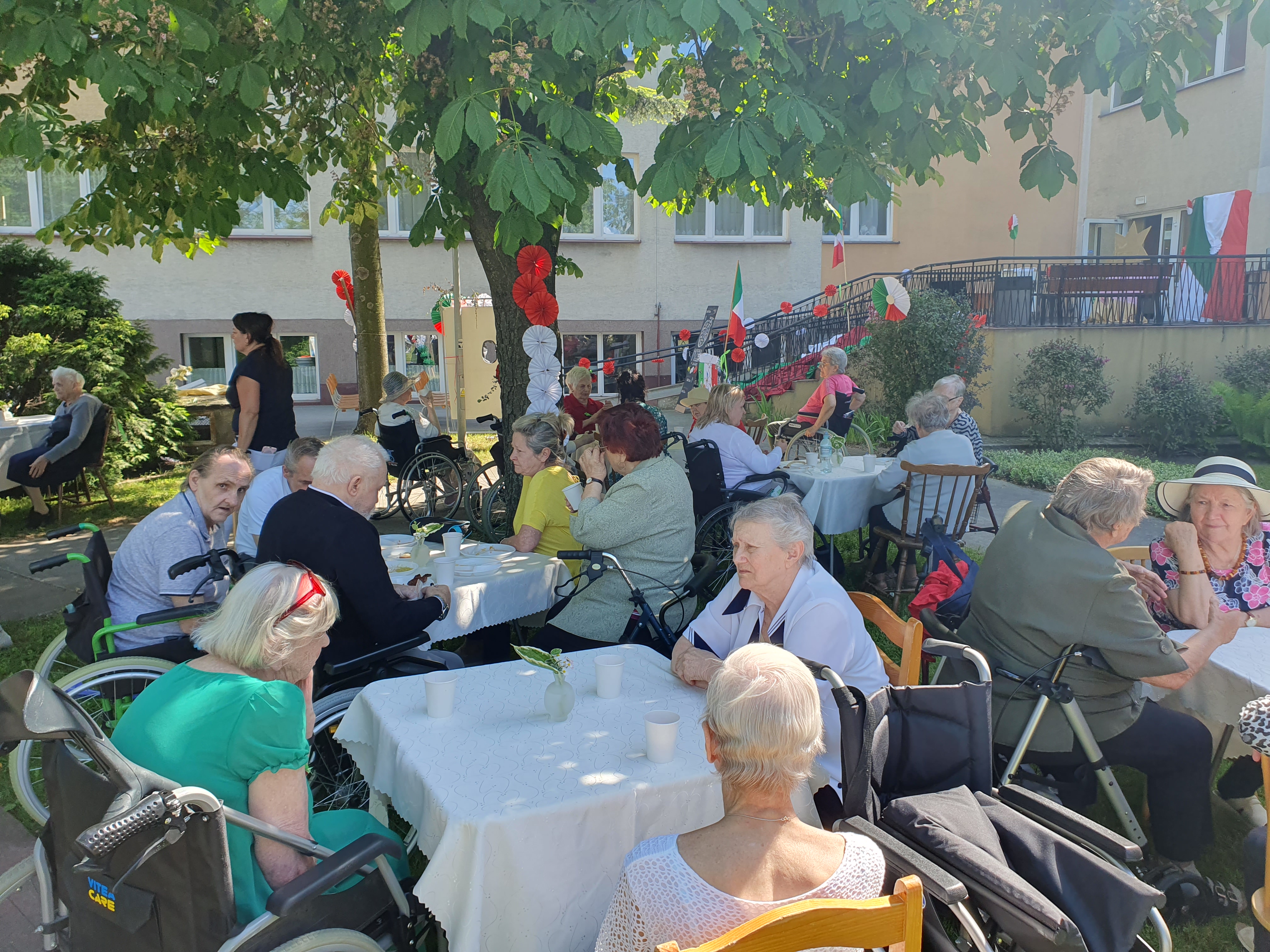 Na ogrodzie DPS znajduje się kilkanaście stołów, przy których siedzą Seniorzy