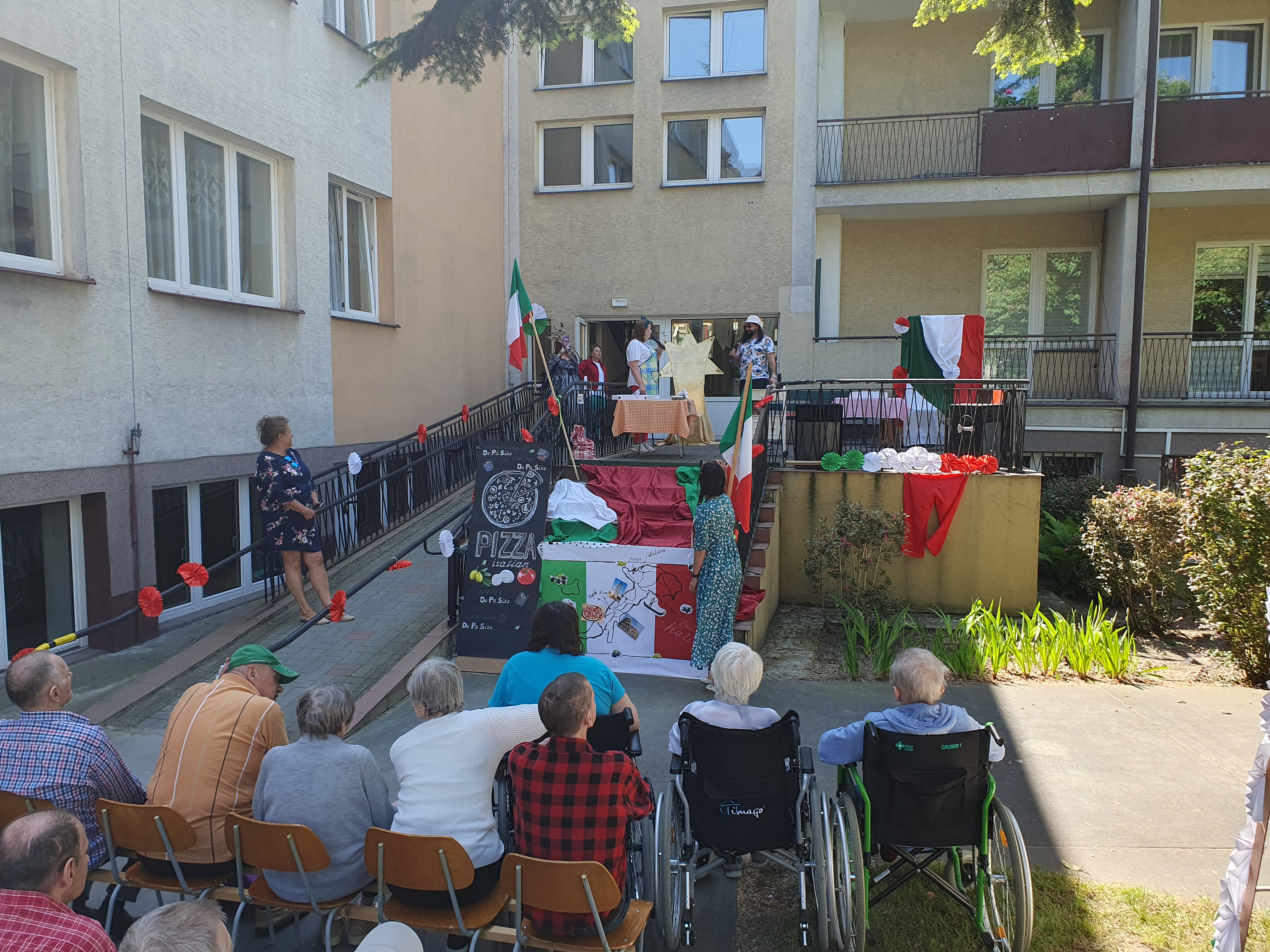 a pierwszym planie znajdują się Seniorzy siedzący na ogrodzie, przed nimi znajdują się schody i wjazd do DPS zaaranżowane we włoskim stylu, z boku znajduje się Pani Dyrektor