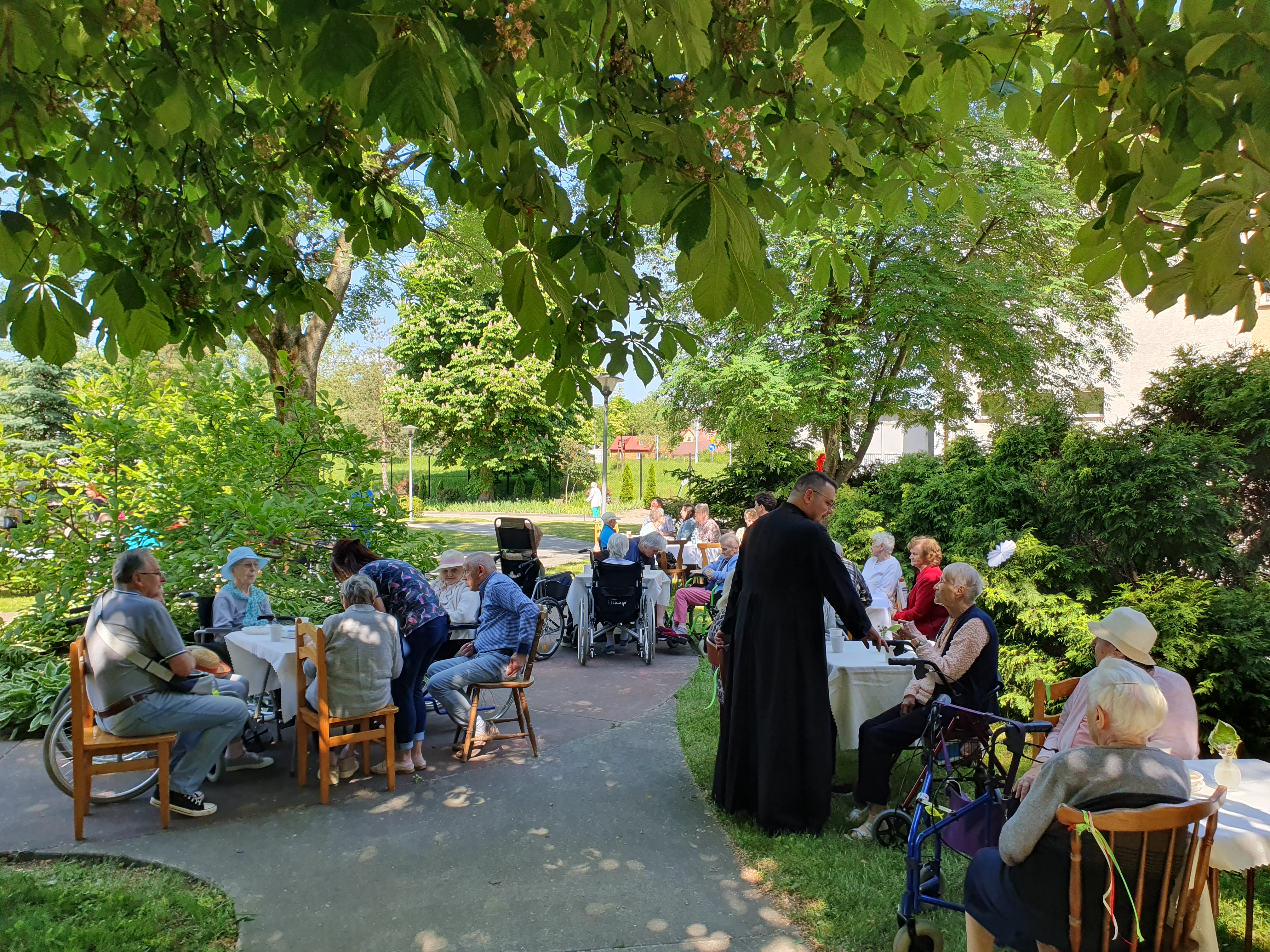 Seniorzy siedzą przy stołach, wśród zieleni ogrodku DPS, przy jednym ze stołów duchowny rozmawia z Seniorami
