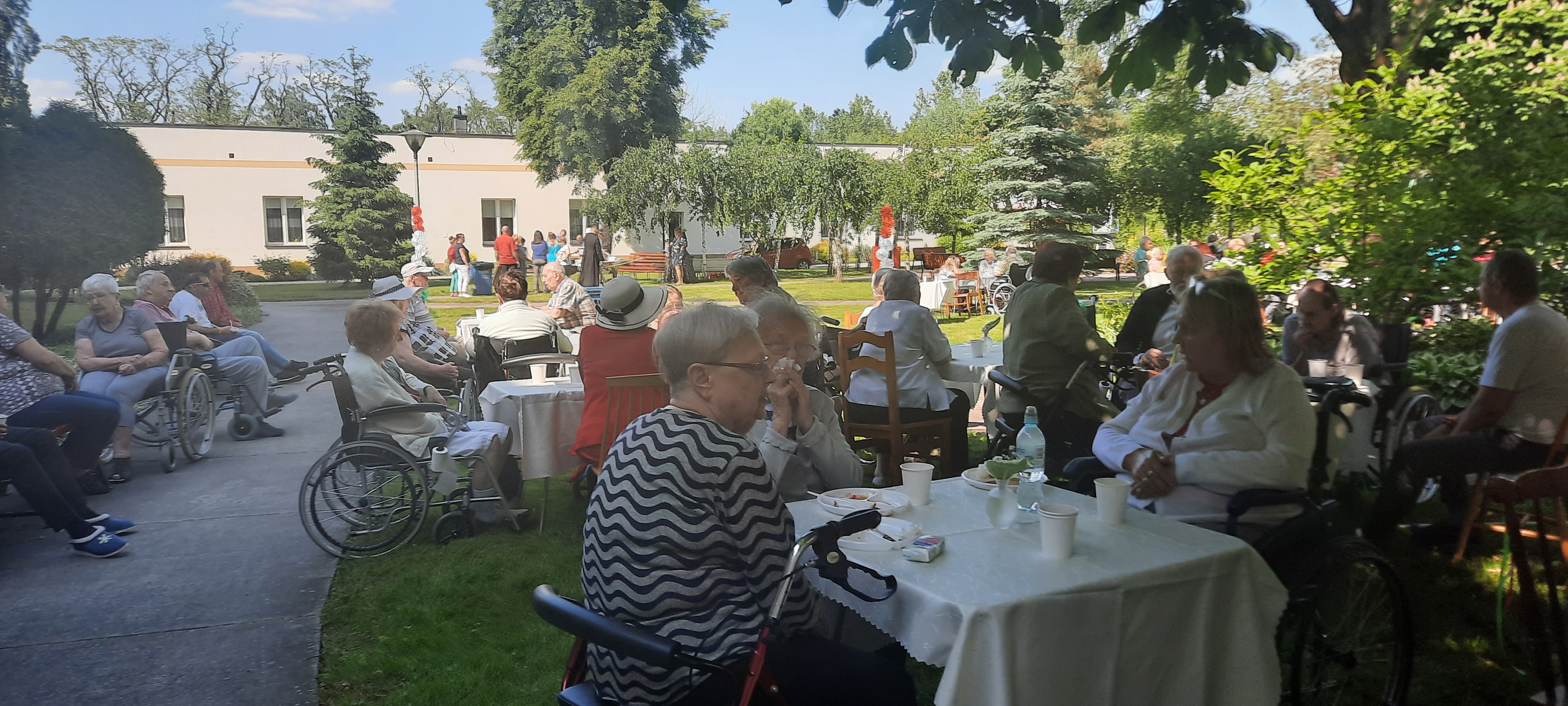 Szerokie ujęcie na imprezę trwającą na ogrodzie dps. W tle widać Seniorów siedzących przy stołach