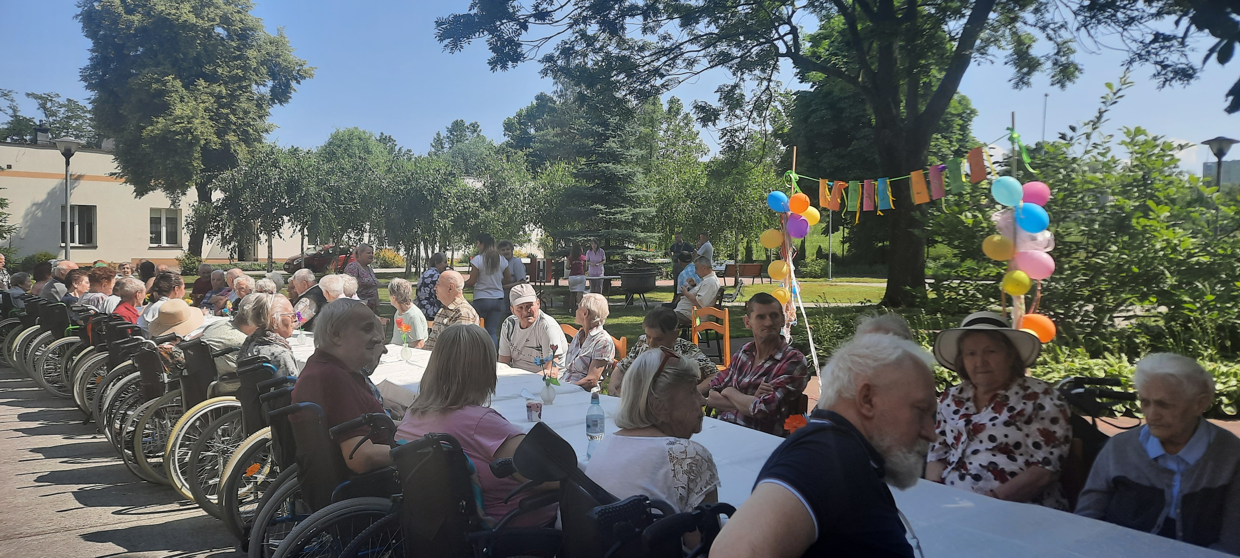 Na ogrodzie DPS przy bardzo długim stole siedzą Seniorzy