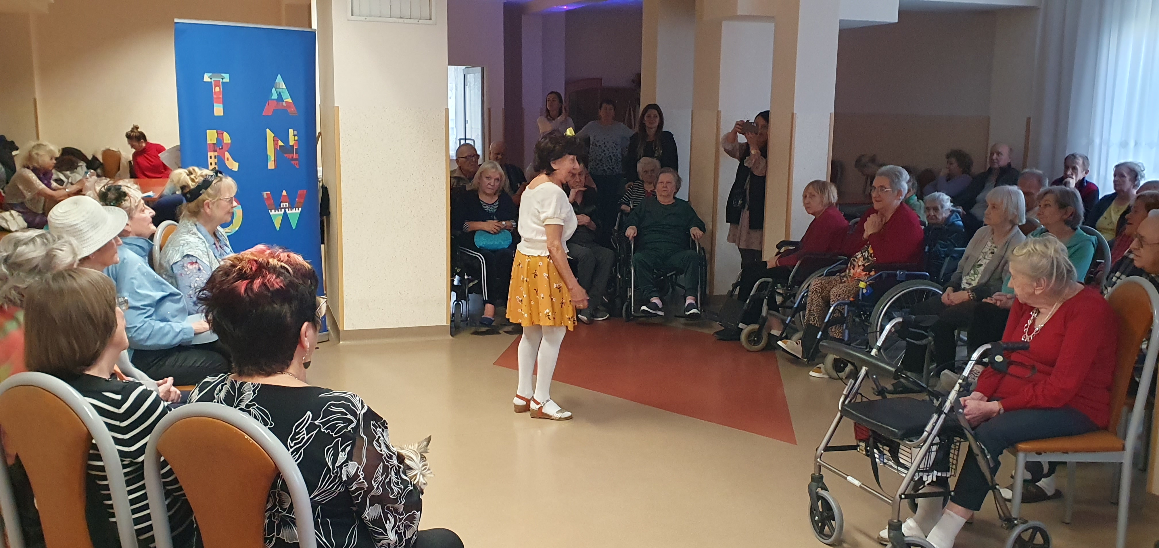 Zdjęcie ze świetlicy domu, grupa Seniorów siedzi dokoła sceny, na której znajduje się Seniorka ubrana w białą bluzkę oraz żółtą spódniczkę