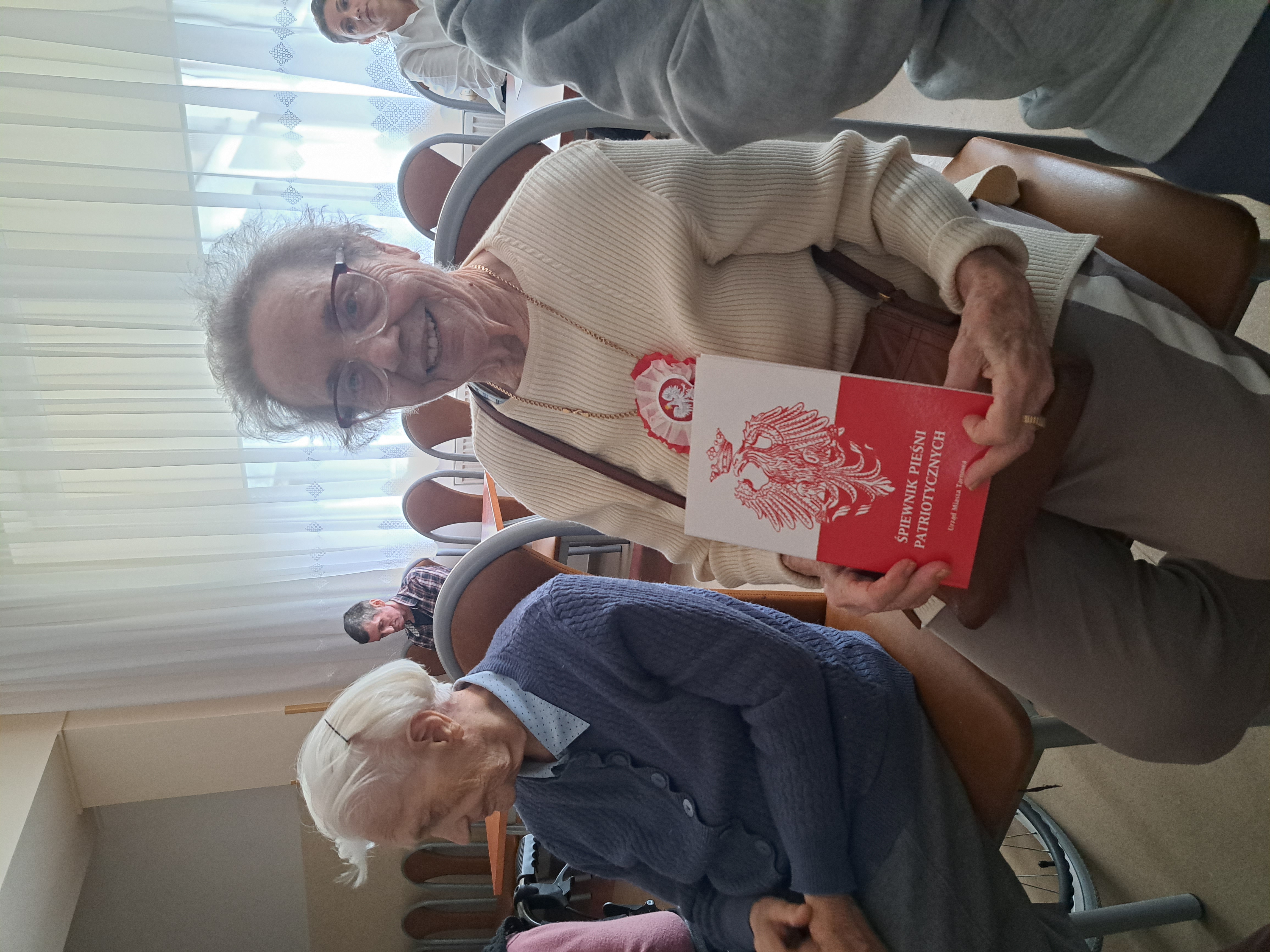 Seniorka pozuje do zdjęcia, siedzi na sali. W ręku trzyma śpiewnik pieśni patriotycznych