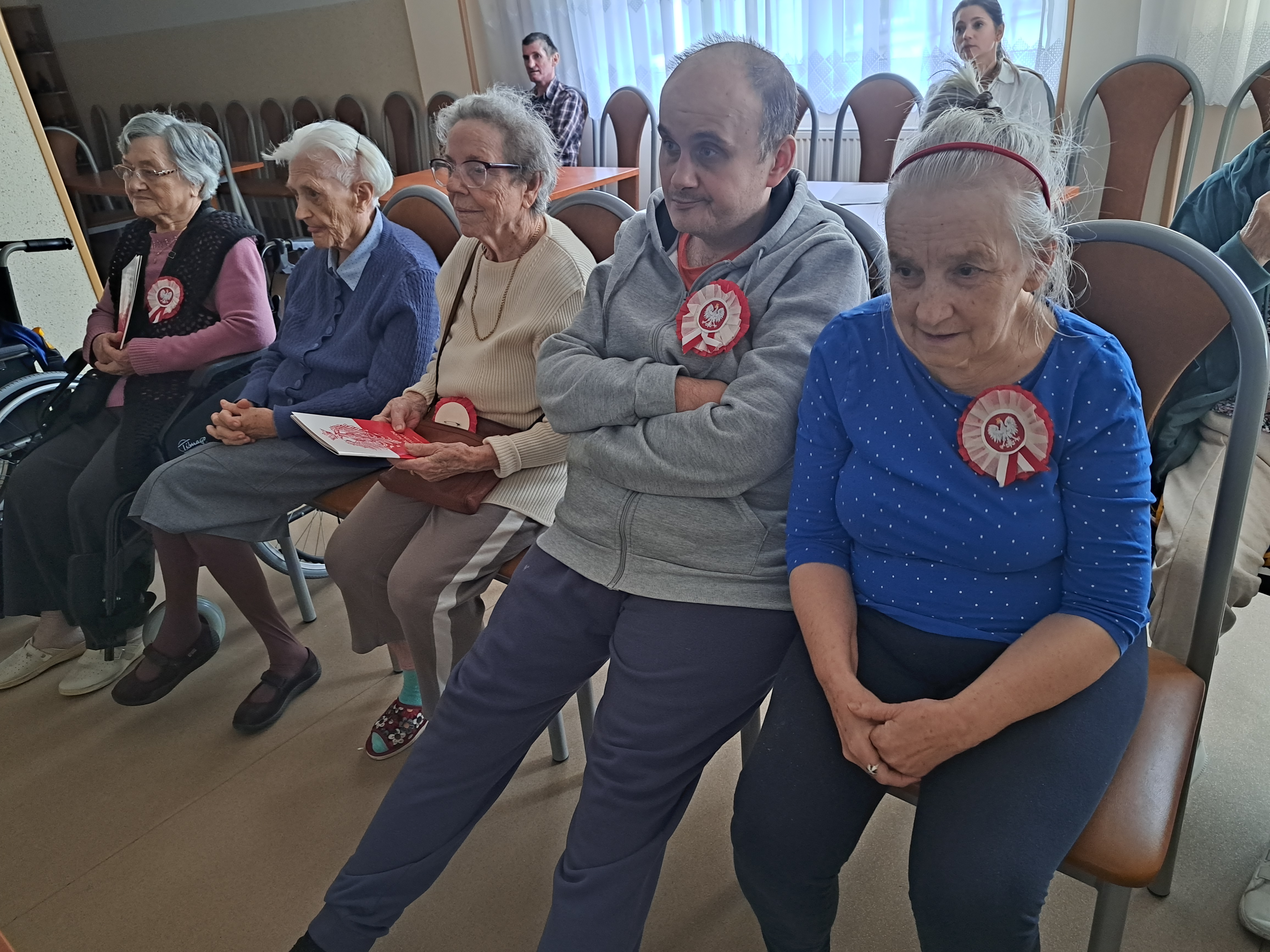 Seniorzy siedzą na sali i śpiewają pieśni patriotyczne