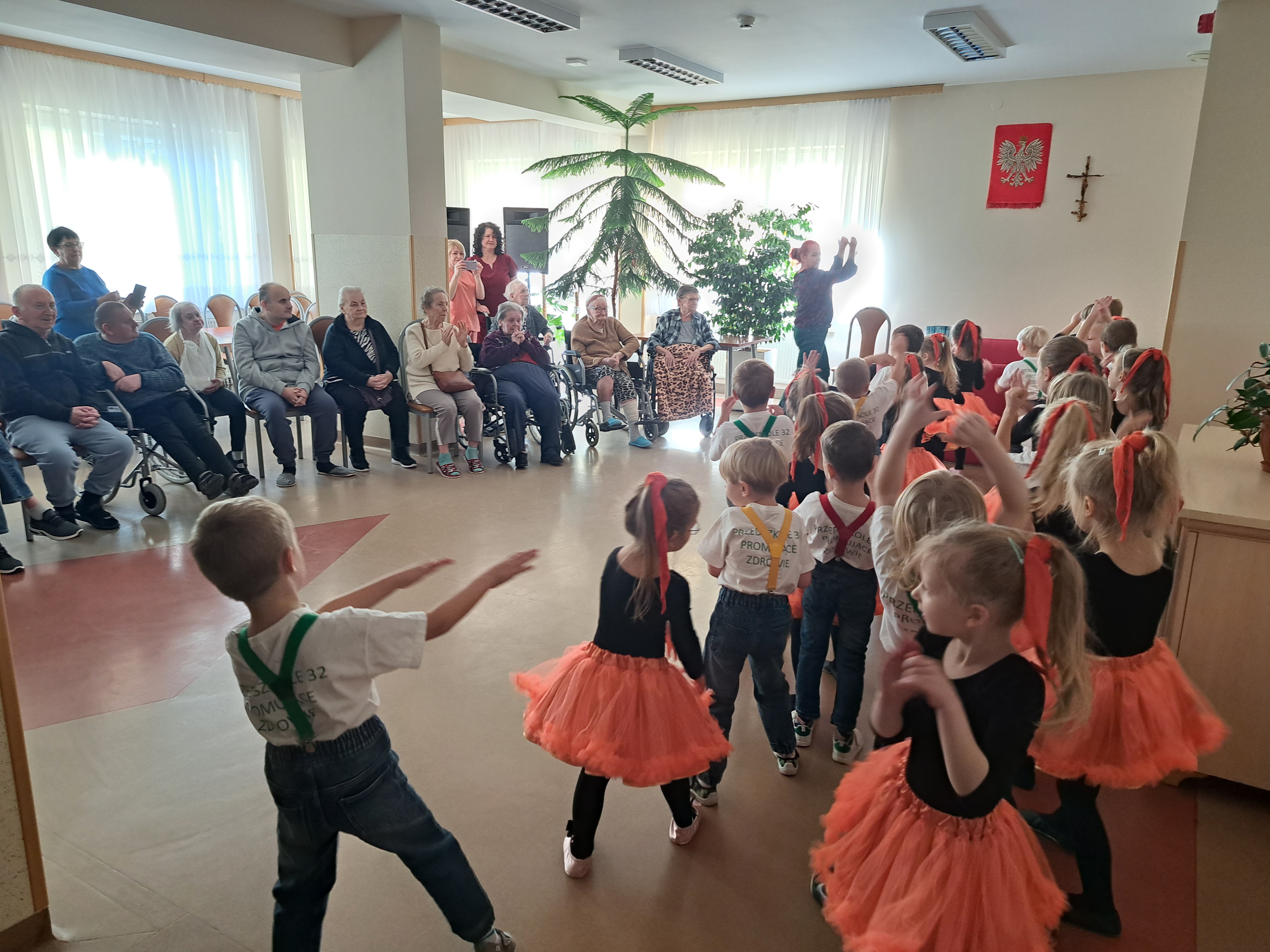 Seniorzy oglądają występ taneczny dzieci, chłopcy są ubrani w czarne spodnie i białe koszulki, a dziewczynki w czarne koszulki i pomarańczowe spódniczki