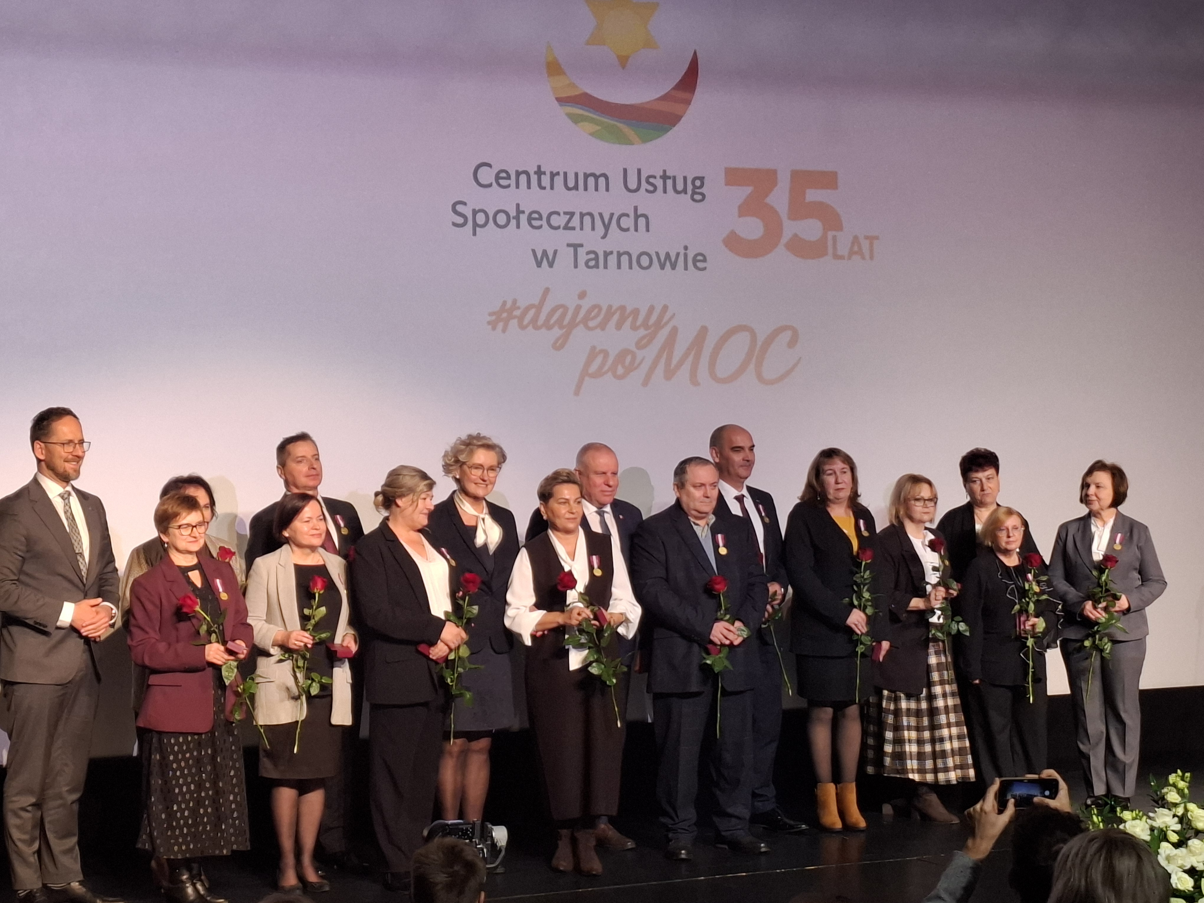 Grupa elegancko ubranych ludzi stoi na scenie, trzymajac róże w ręku, za nimi znajduje wyświetlony napis Centrum Usług Wspólnych w Tarnowie 35 lat dajemy PoMOC