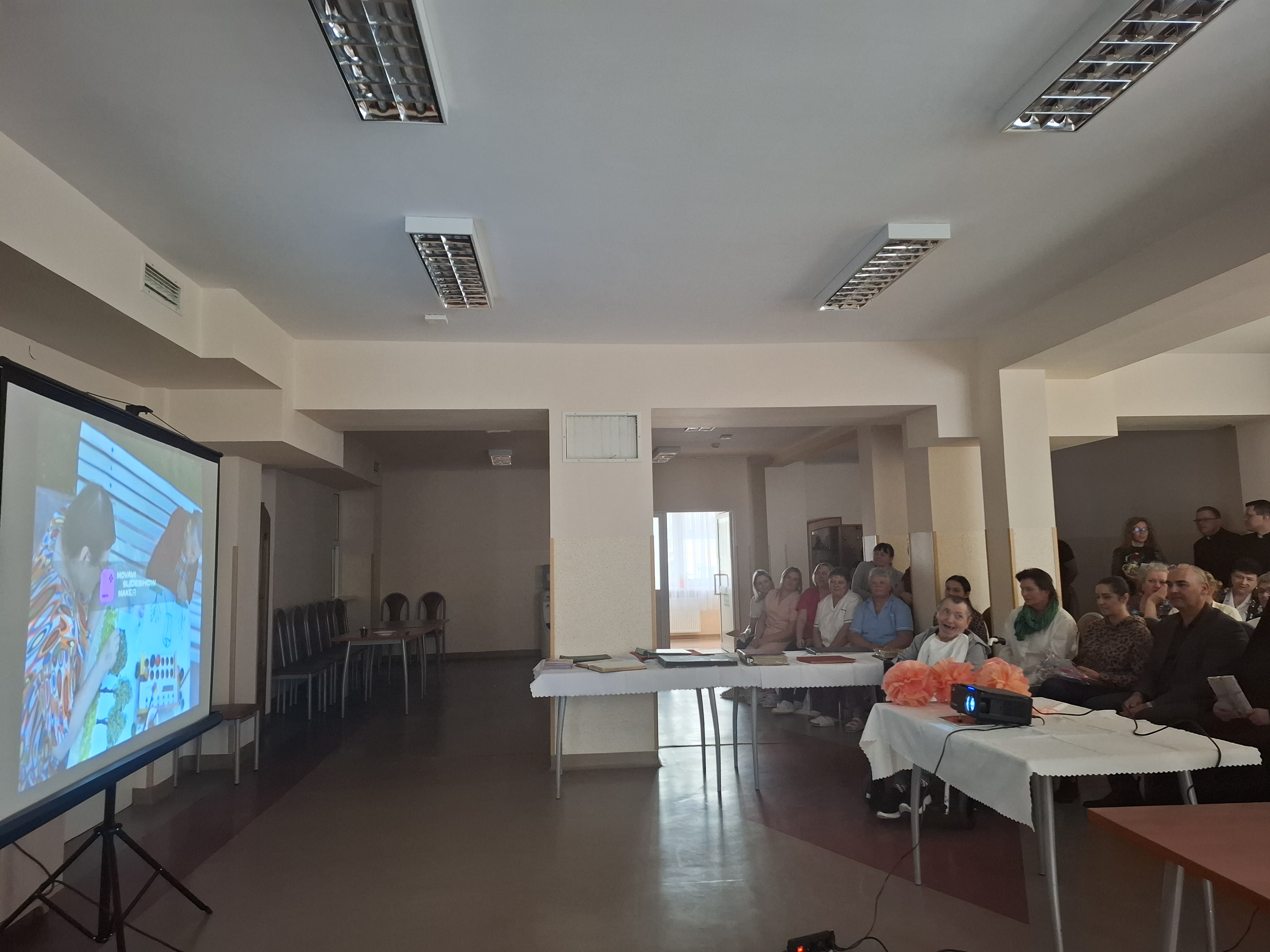 Ujęcie na świetlicę Domu, przed wyświetloną na rzutniku prezentacją siedzą Seniorzy oraz Pracownicy