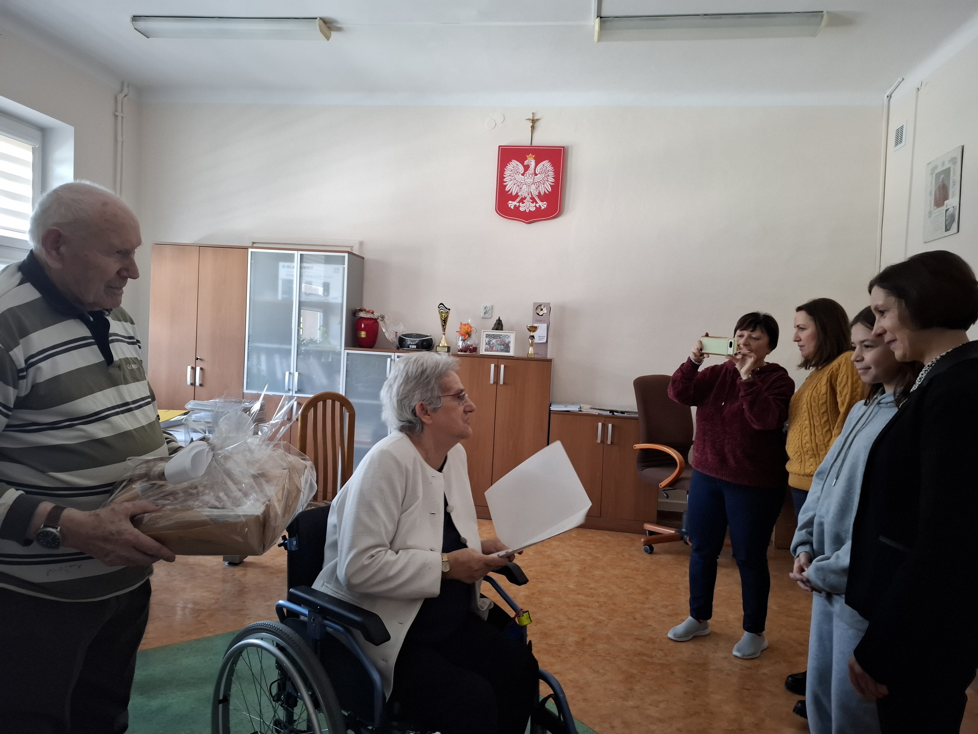 Senior stoi z koszem ciastek, przed nim znajduje się Seniorka na wózku, mówiąca do stojącej przed nimi młodzieży