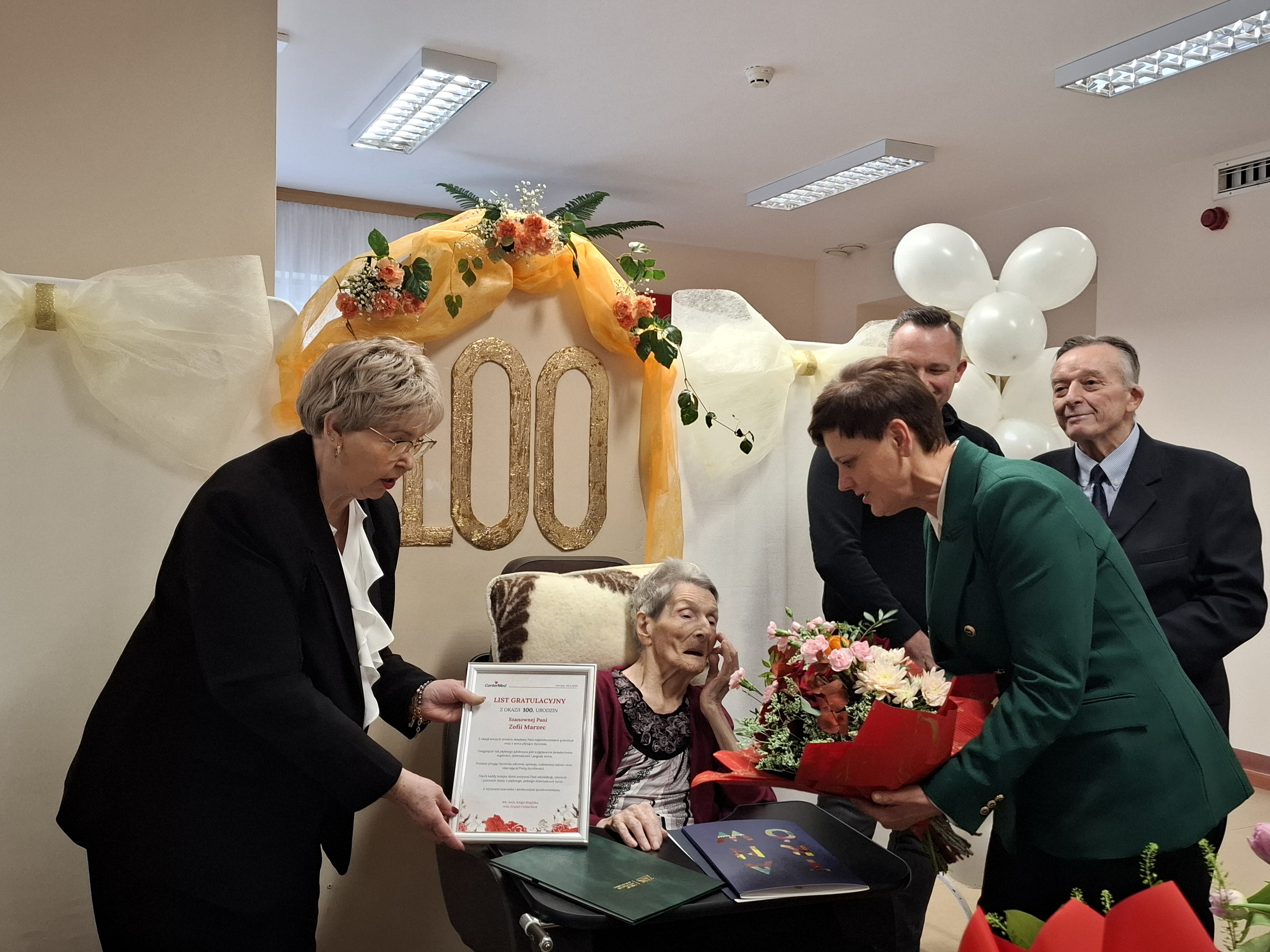 Pani Zofia otzymuje bukiet kwiatów oraz list gratulacyjny z okazji setnych urodzin