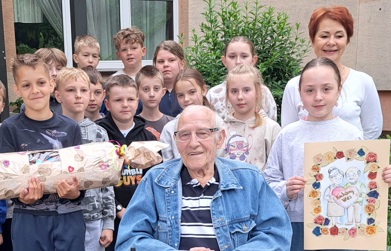 Wspólne zdjęcie Seniora oraz dzieci ze szkoły podstawowej na tle placówki szkolnej
