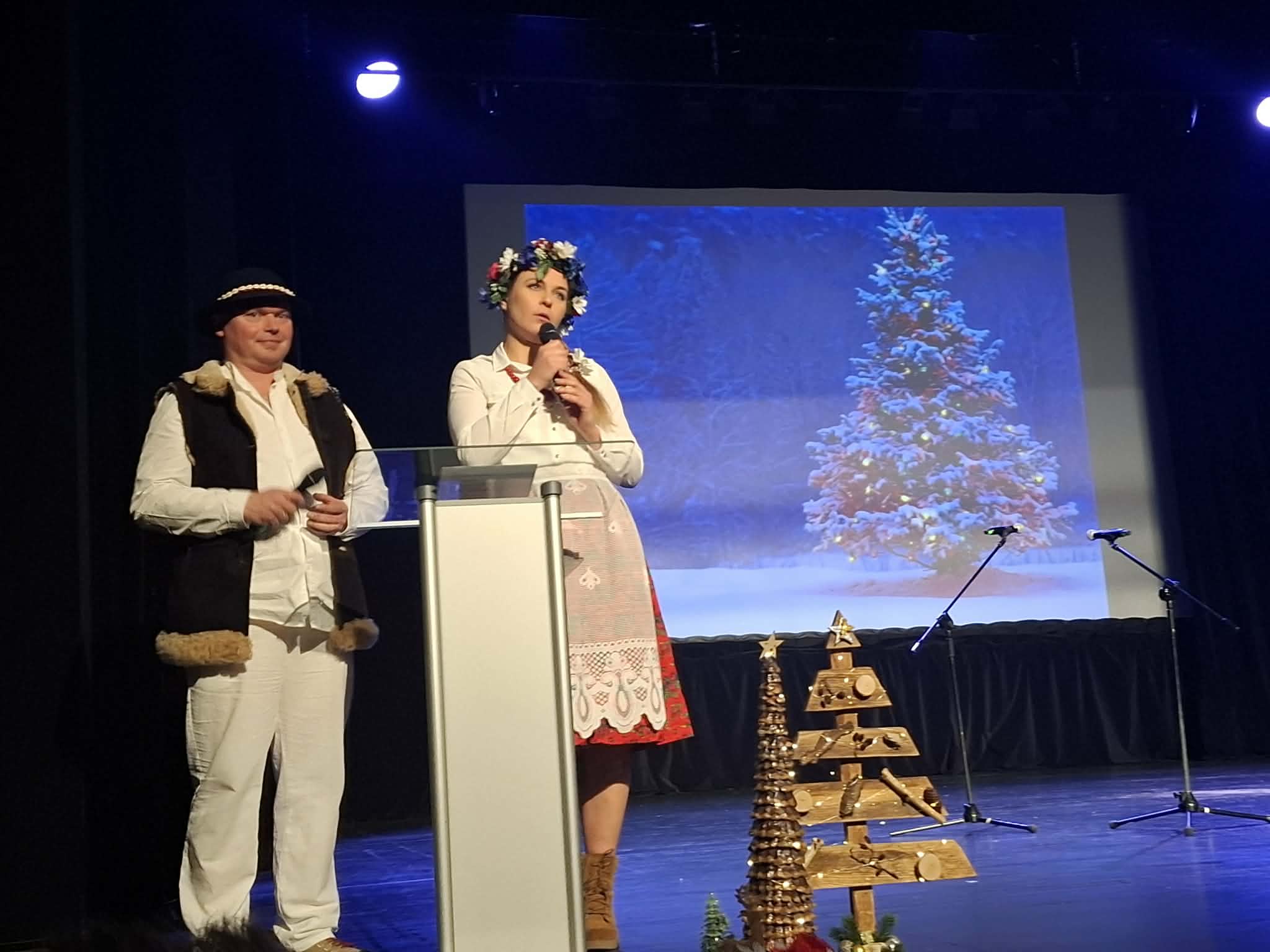 Kobieta w ludowym stroju oraz mężczyzna w góralskim stroju stoją na scenie, obok nich jest rzutnik wyświetlający świąteczne drzewko