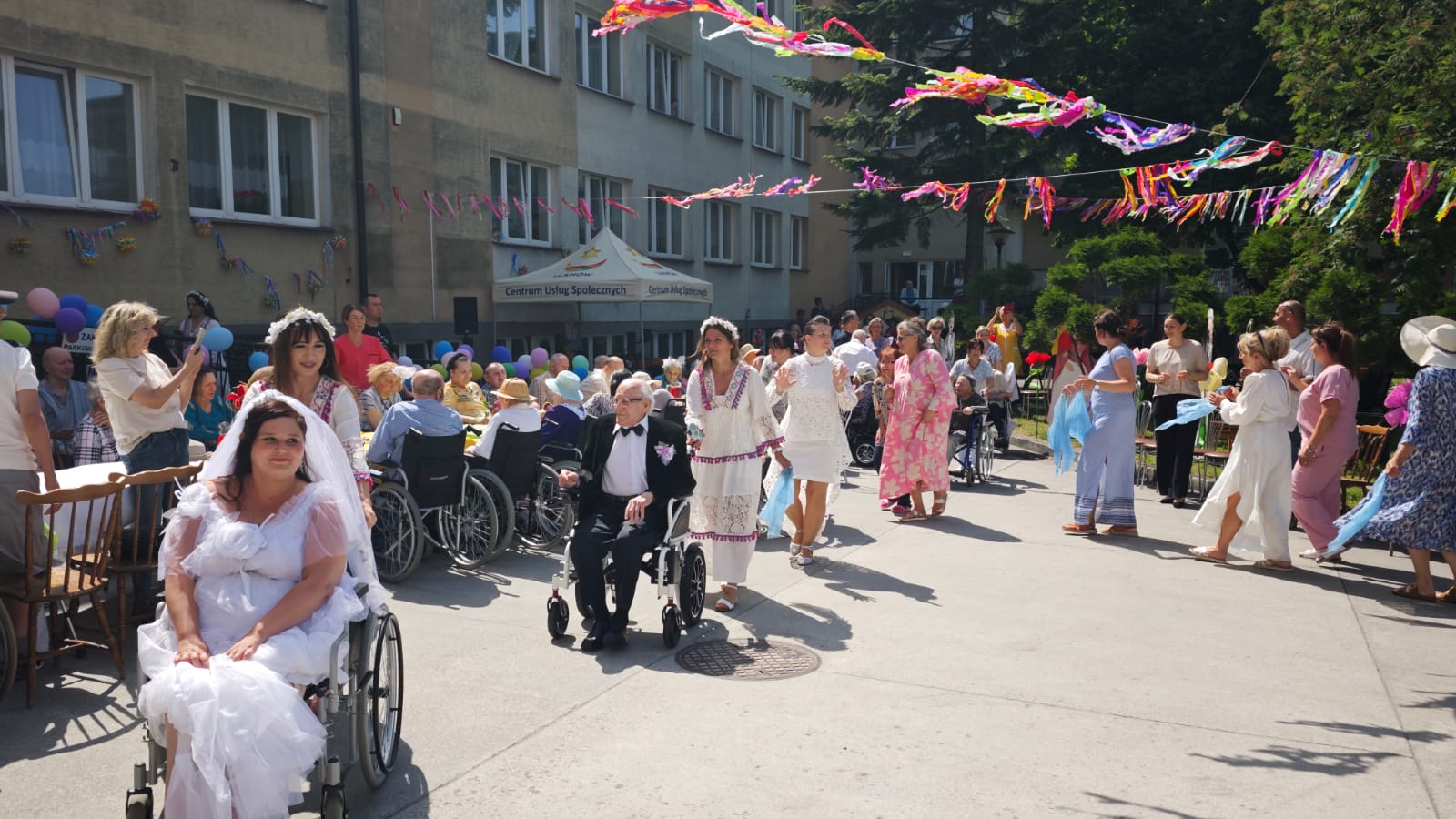 Mieszkańcy oraz pracownicy Domu tańczą na ogródku Domu, na pierszym planie kobieta na wózku inwalidzkim ubrana w sukinie ślubną, a za nią Senior ubrany w garnitur