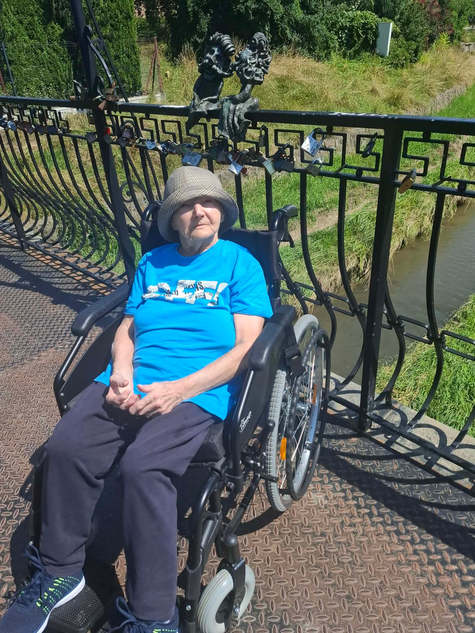 Seniorka na wózku inwalidzkim pozuje do zdjęcia na mostku, który jest ozdobiony rzeźbą Maszkaronów Zakochanych