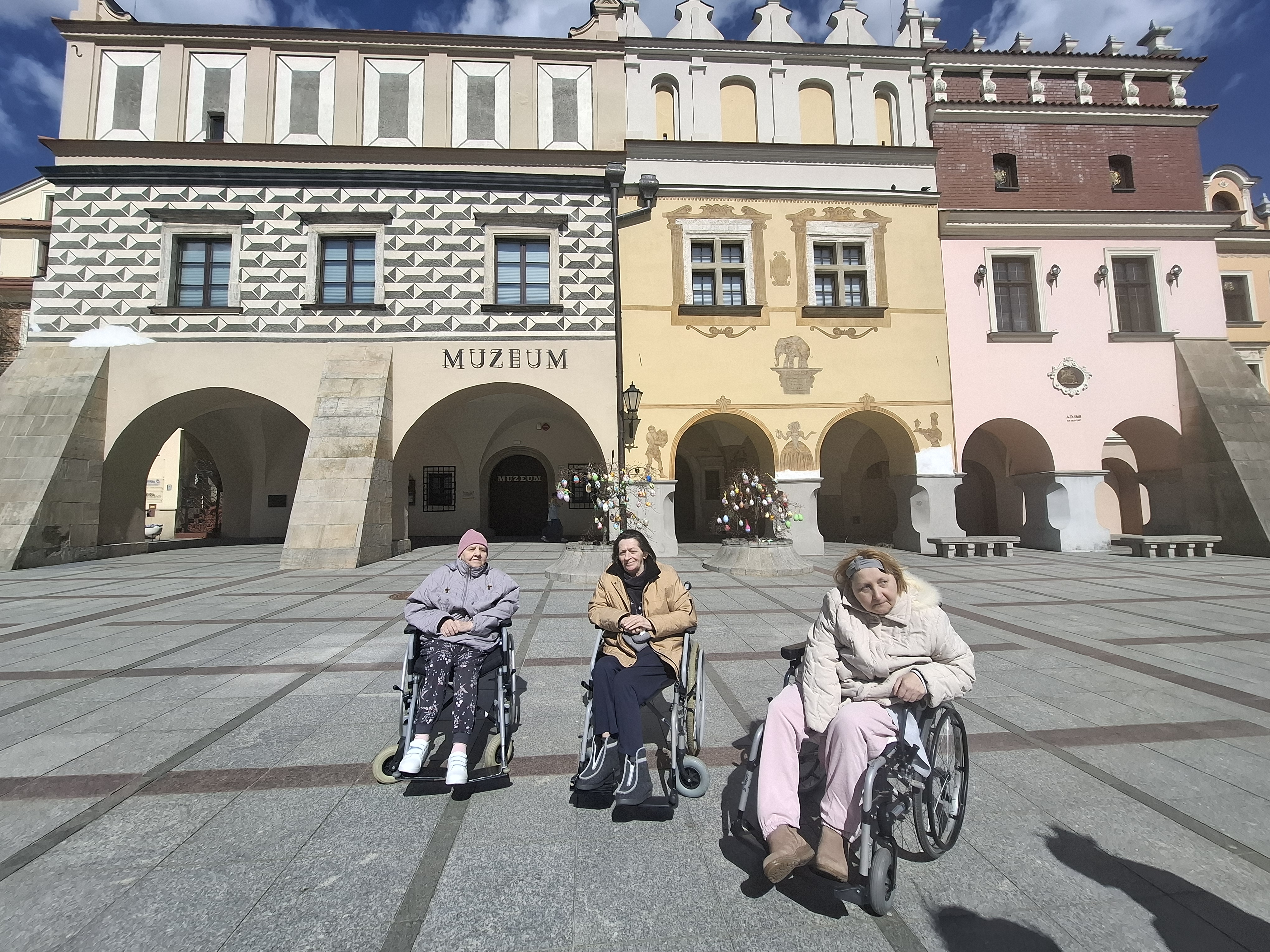Trzy Seniorki na wózkach inwalidzkich pozują do zdjęcia na tle tarnowskiego muzeum znajdującego się na Rynku