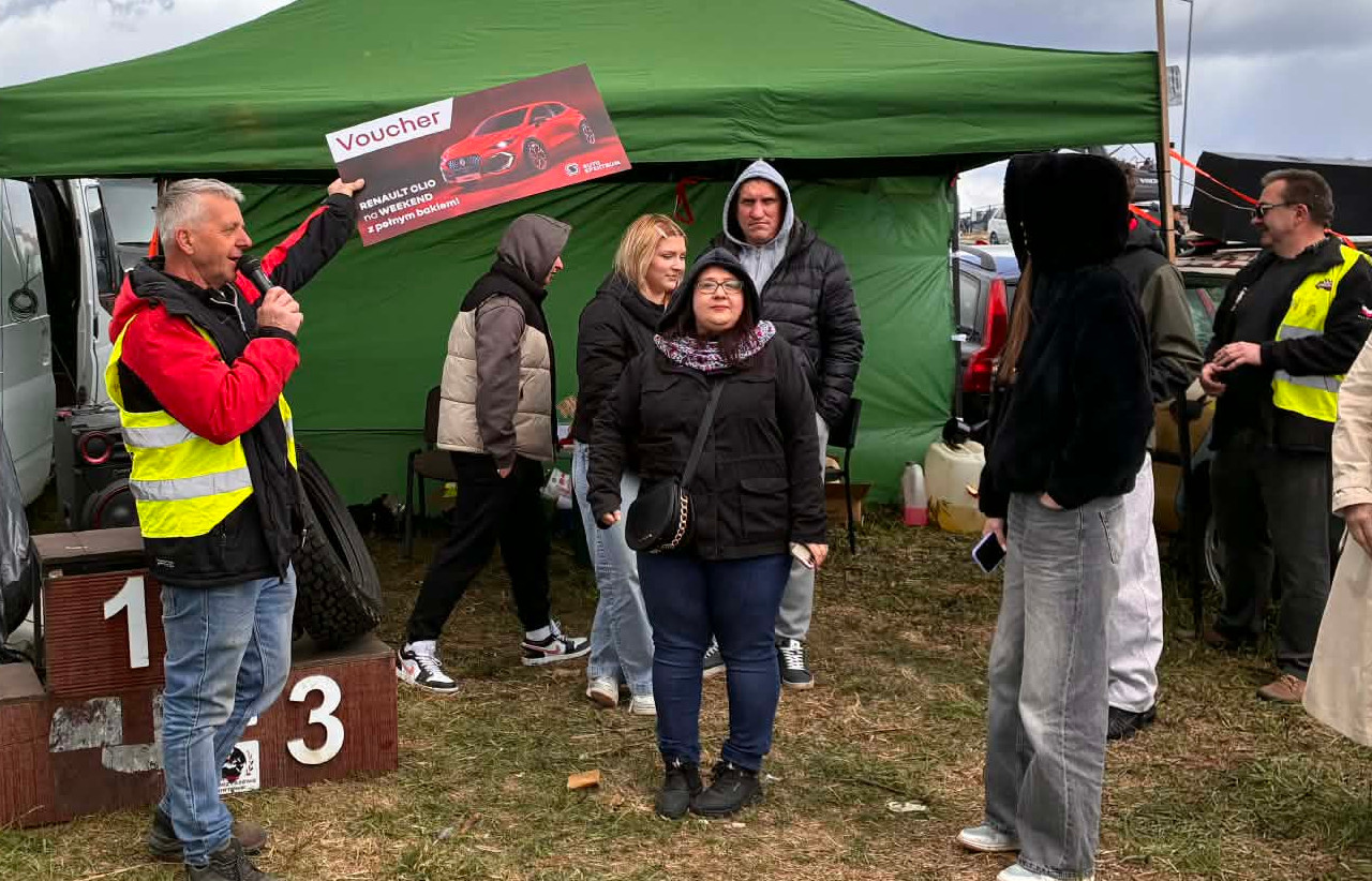 Prowadzący event Wrak Race Tarnów stoi z mikrofonem trzymając w rękach plakat wydarzenia, wokól stoją uczestnicy wydarzenia