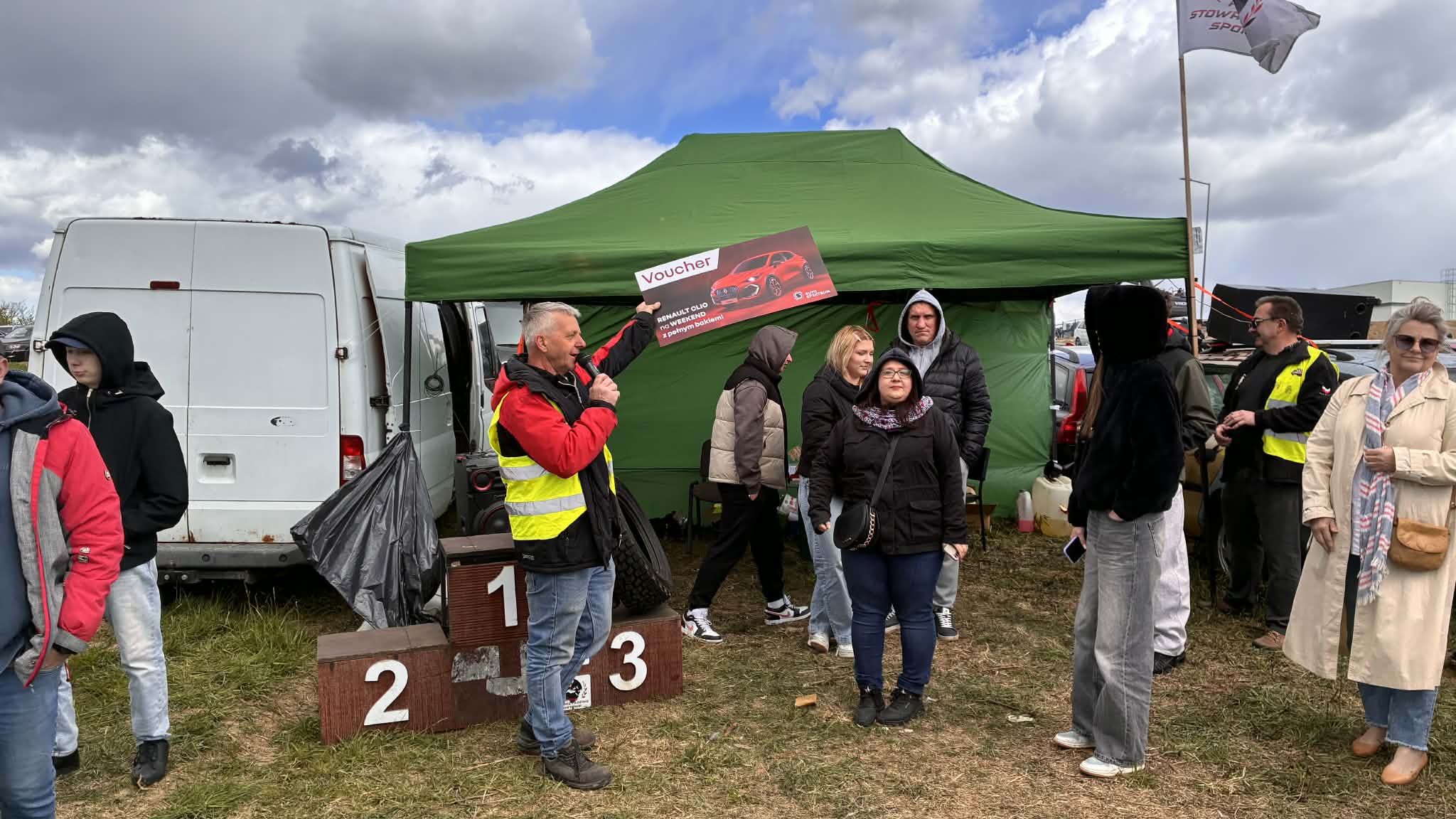 Prowadzący event stoi z mikrofonem w ręku oraz plakatem wydarzenia, w tle znajduje się zielony namiot oraz podium z trzema miejscami, dokoła stoją uczestnict wydarzenia